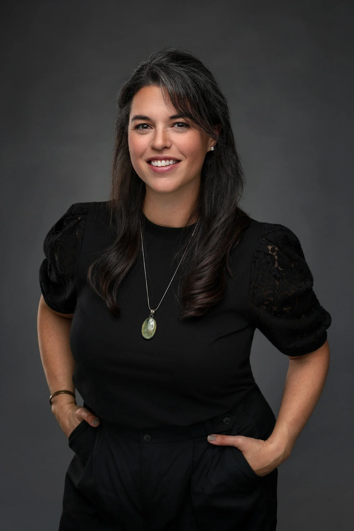 A woman with dark hair and light skin, wearing a black top with lace sleeves, standing against a dark gray background, smiling, with hands in her pockets