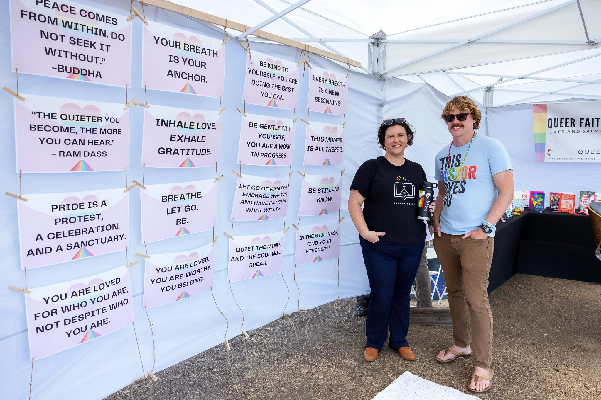 Two women standing inside a white canopy tent with various printed inspirational quotes hanging on the back wall. One woman is in black holding a drink, and the other is in a light blue shirt with colorful text, both smiling at the camera.