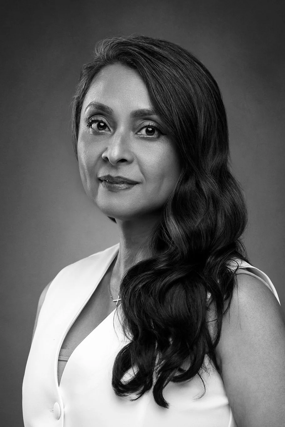 Black and white portrait of a woman with long wavy hair, wearing a sleeveless white top and a necklace, looking at the camera