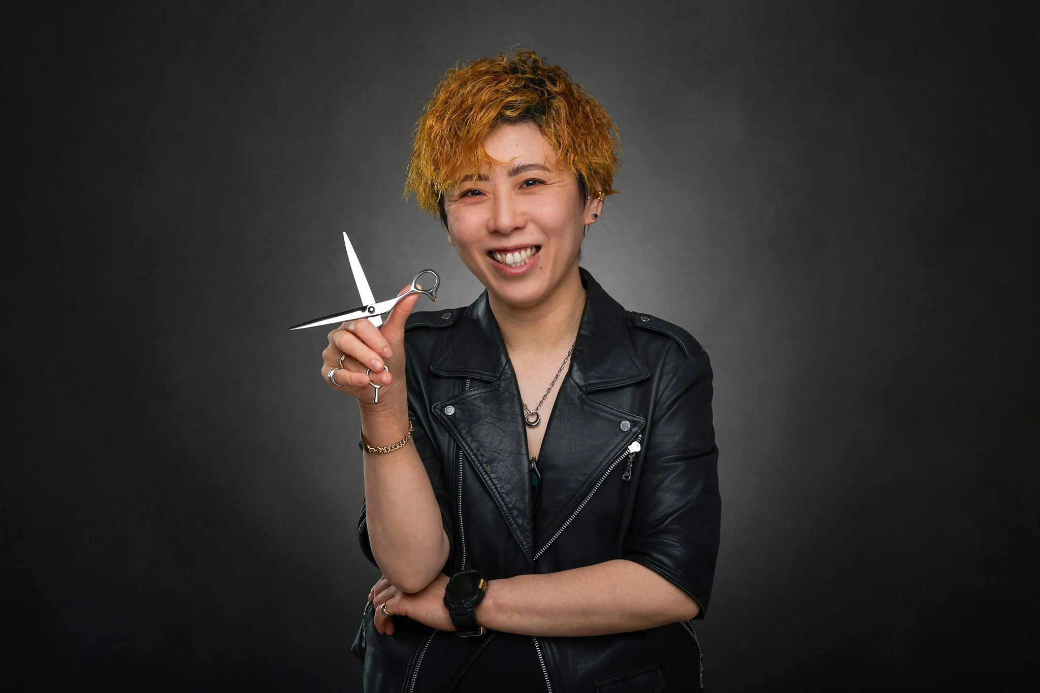A person with short, curly blond hair styled in an asymmetrical cut, smiling and holding a pair of scissors near their head. They are wearing a black leather jacket, multiple rings, and a black watch, against a dark background.