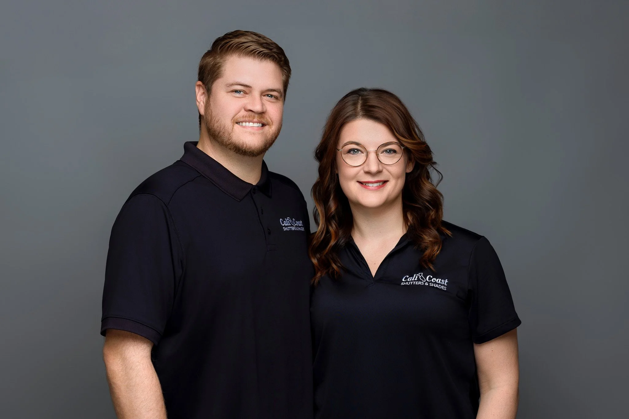 A man and a woman posing together against a plain gray background. Both are wearing black shirts with a logo that reads "Cali Coast Shutters & Shades." The man has light brown hair, a beard, and is smiling, while the woman has long, wavy brown hair, 