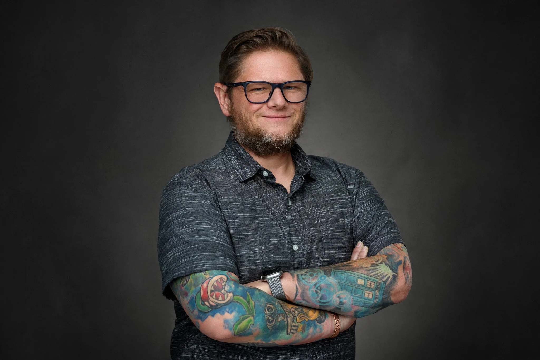 A smiling man with glasses, a beard, and tattoos on his arms, standing with crossed arms against a dark background.