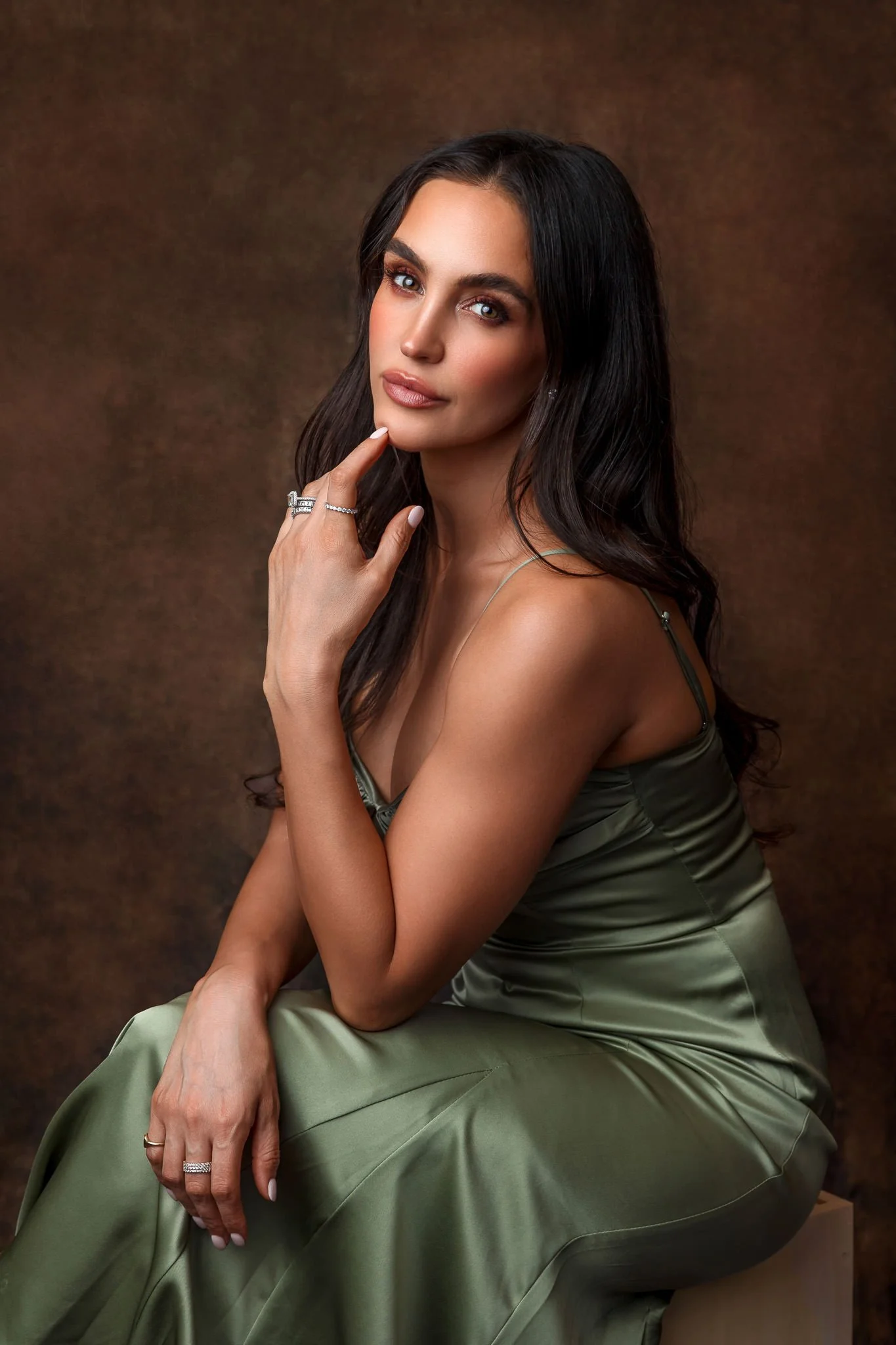 A woman with long dark hair and blue eyes, wearing a satin green dress with thin straps, sitting against a brown background.