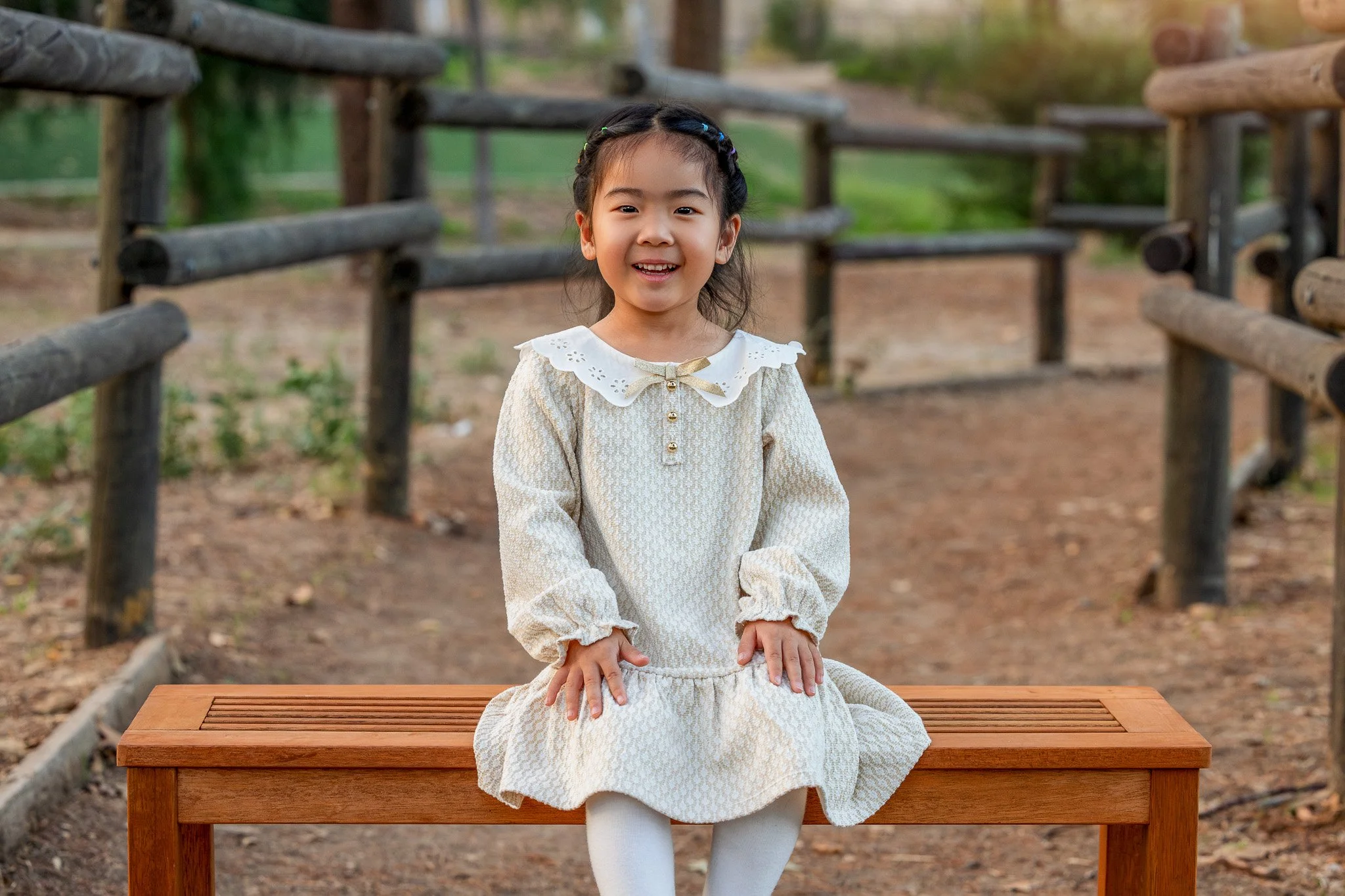 Young girl with dark hair and a braid, smiling, sitting on a wooden bench outdoors with a dirt path and wooden railing in the background, wearing a cream-colored dress with a white collar and white tights.
