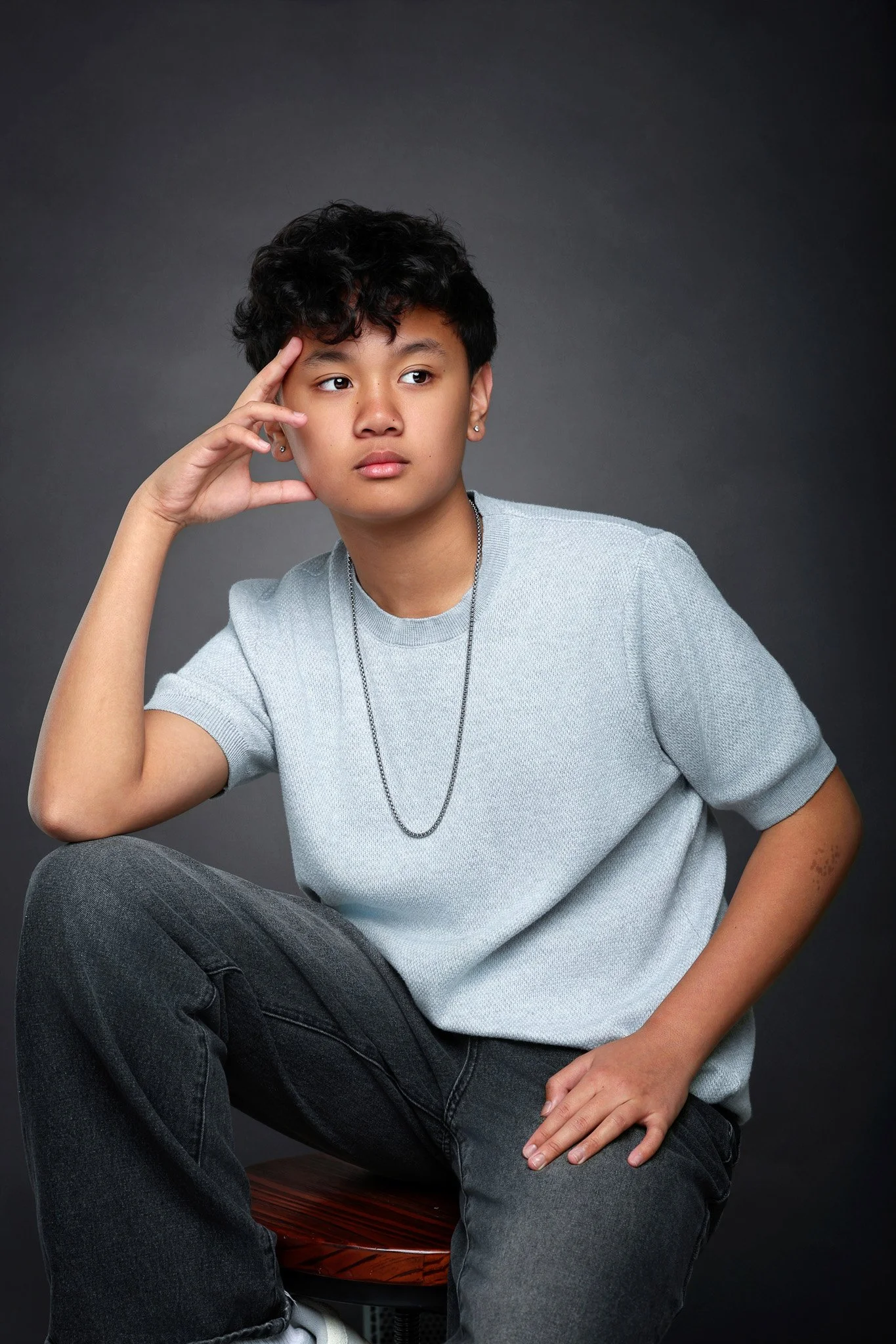 A young person with short, curly black hair, wearing a light gray T-shirt and dark jeans, sitting on a wooden stool with one hand resting on their knee and the other hand touching their temple, looking thoughtfully to the right against a dark gray ba