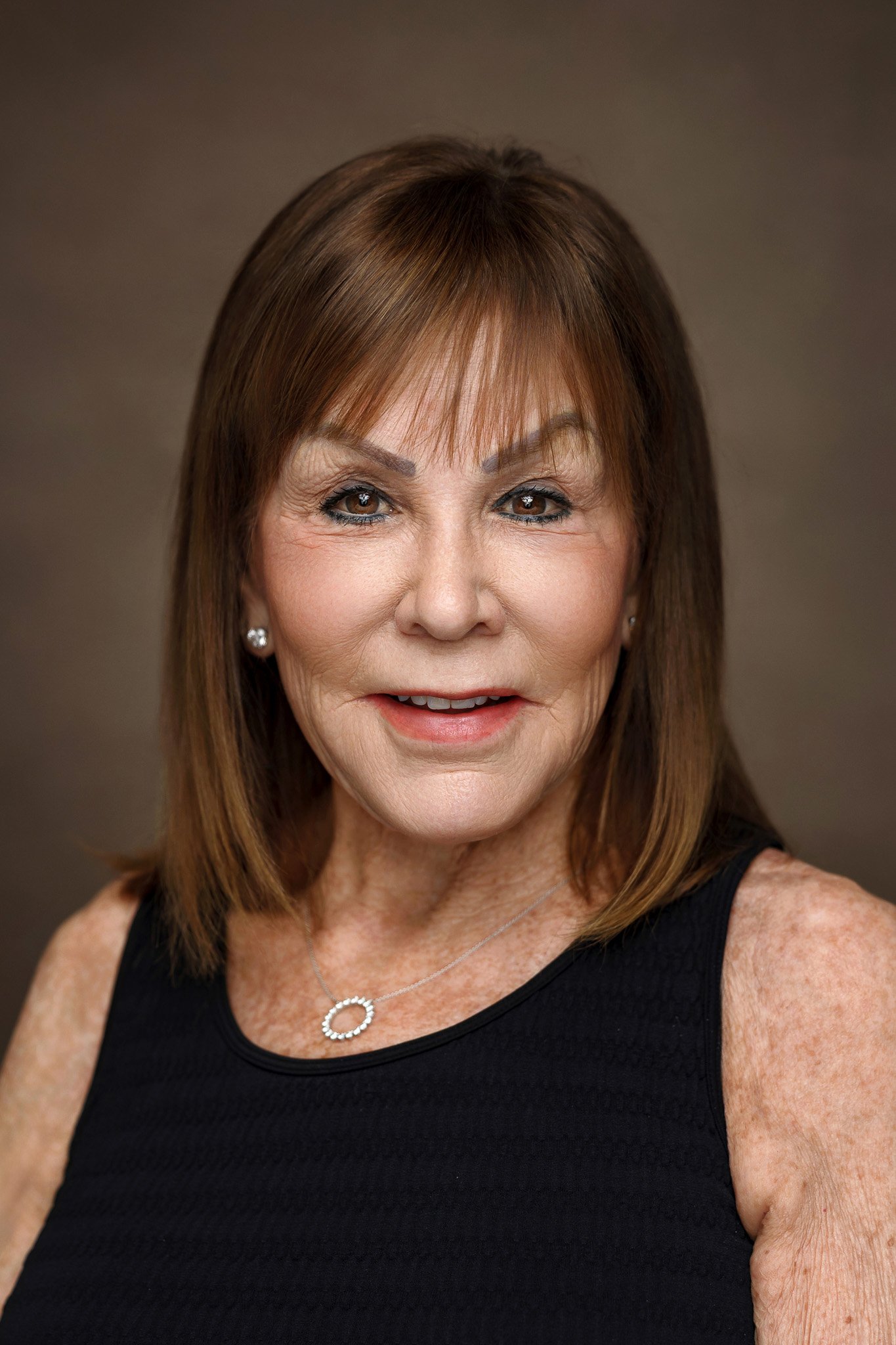 A middle-aged woman with light skin, shoulder-length brown hair, and blue eyes, wearing a black sleeveless top, pearl earrings, and a circular necklace, poses for a portrait against a neutral background.