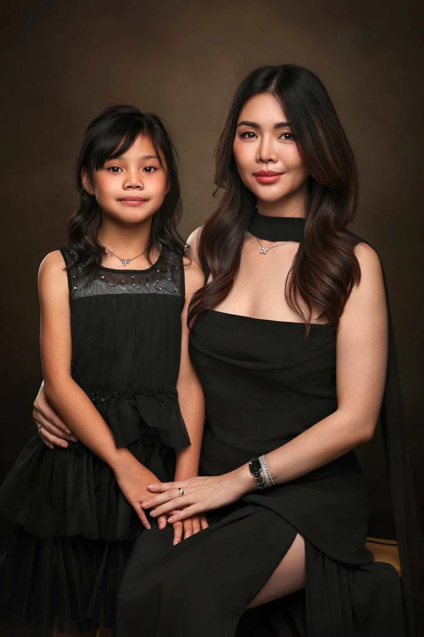 A woman and a young girl dressed in black, sitting and posing together against a dark brown background. They are both wearing jewelry and have long dark hair.