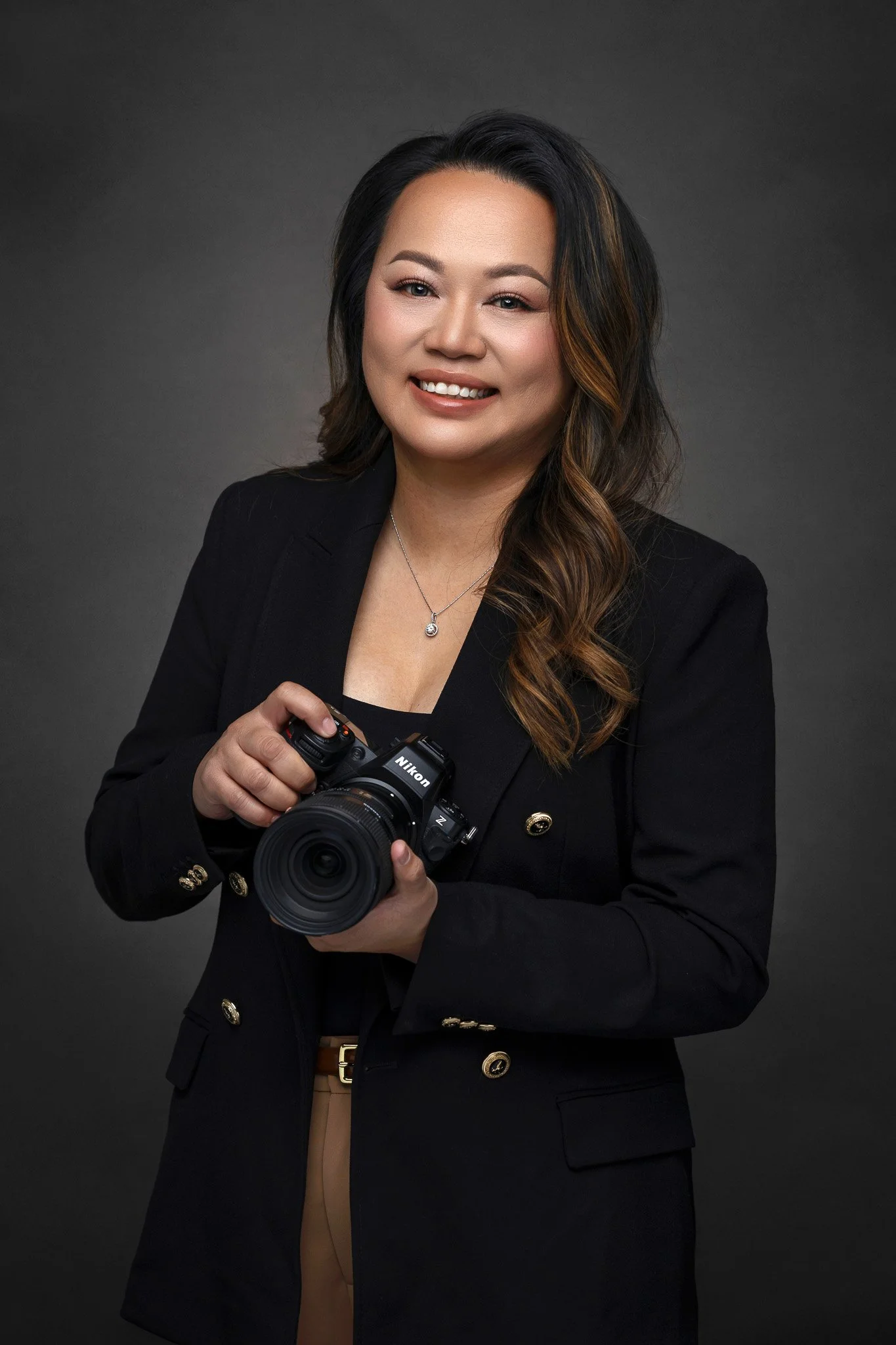 A woman with long wavy hair, wearing a black blazer and holding a Nikon camera, smiling at the camera.
