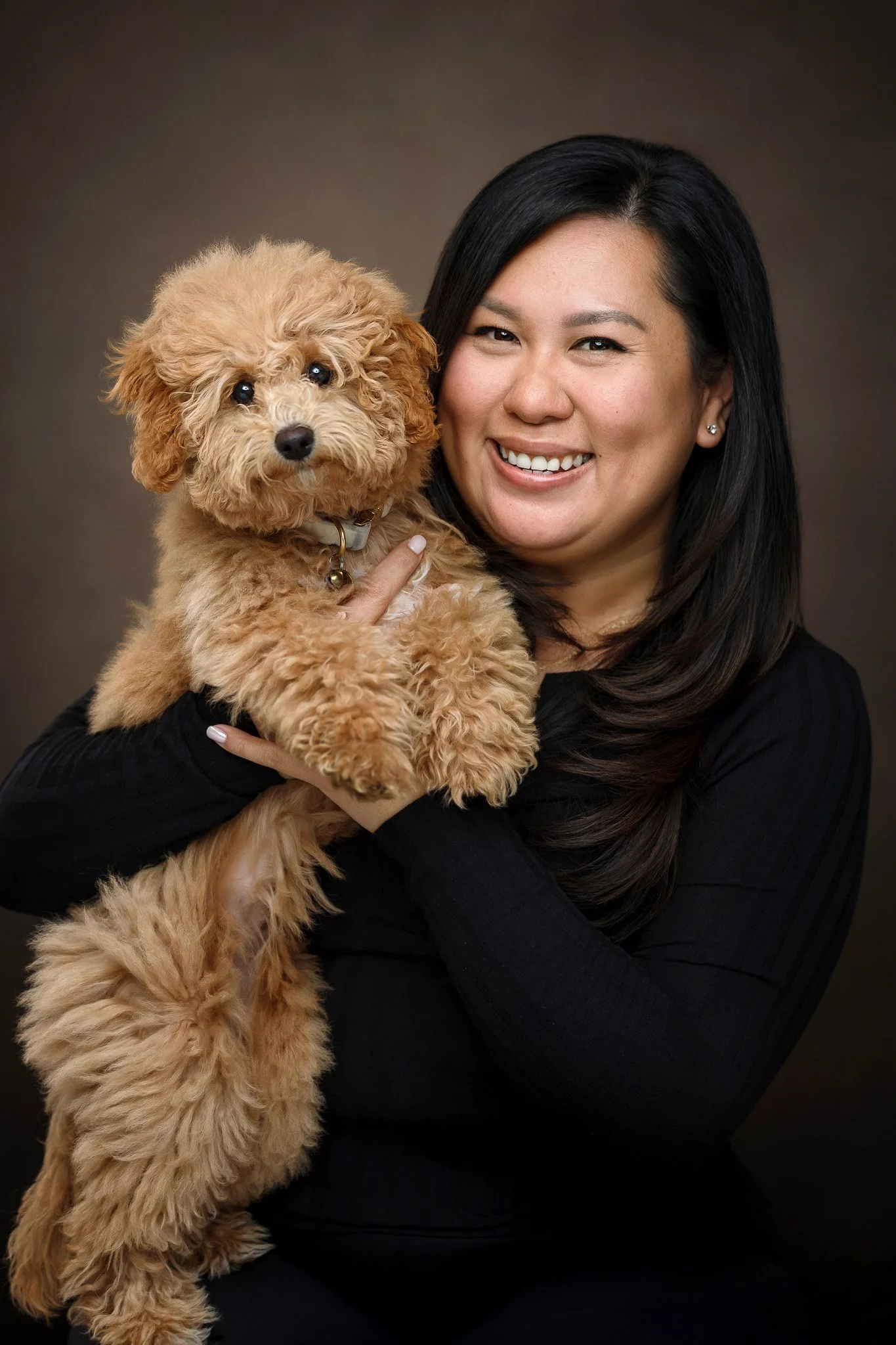 A woman with dark hair smiling while holding a small fluffy tan dog.