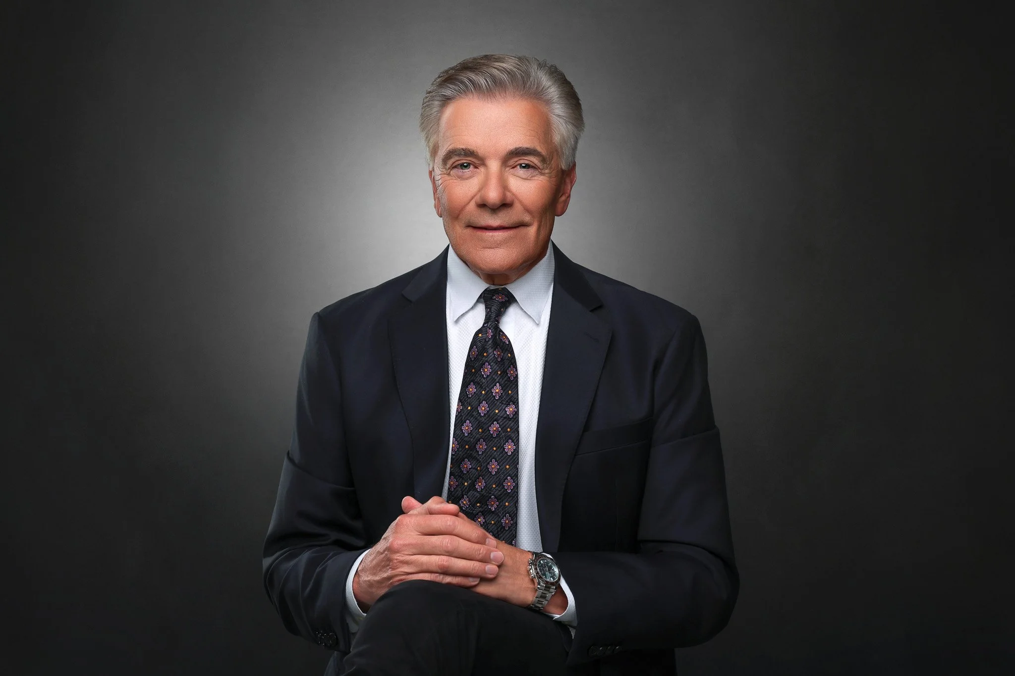 A senior man in a dark suit and tie, with gray hair, smiling, and holding his hands together in front of him against a dark gray background.