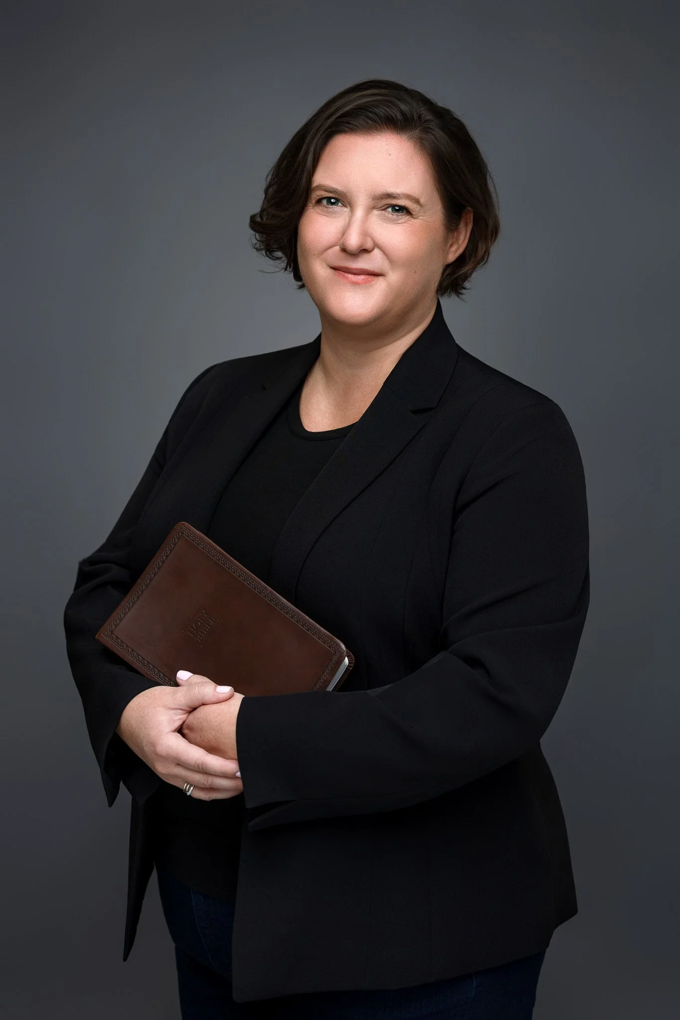 A woman with short dark hair wearing a black blazer and black shirt, holding a brown leather-bound journal, standing against a plain gray background.