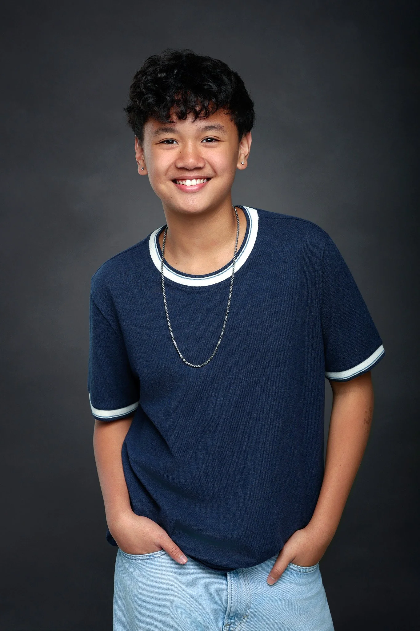 Young person with short curly hair smiling, wearing earrings, a navy blue t-shirt with white trim, light blue jeans, and silver jewelry, standing against a dark background.