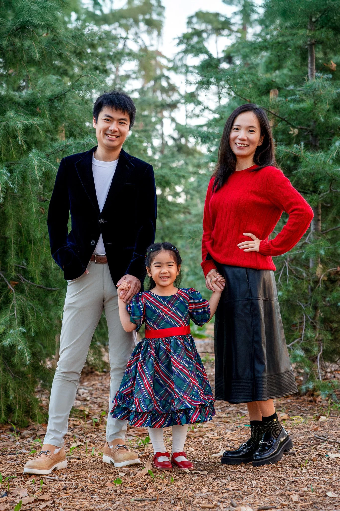 A smiling family of three standing outdoors in a forest, holding hands. The father, on the left, is wearing a black blazer and beige pants. The mother, on the right, is wearing a red sweater and a black skirt. Their young daughter, in the middle, is 