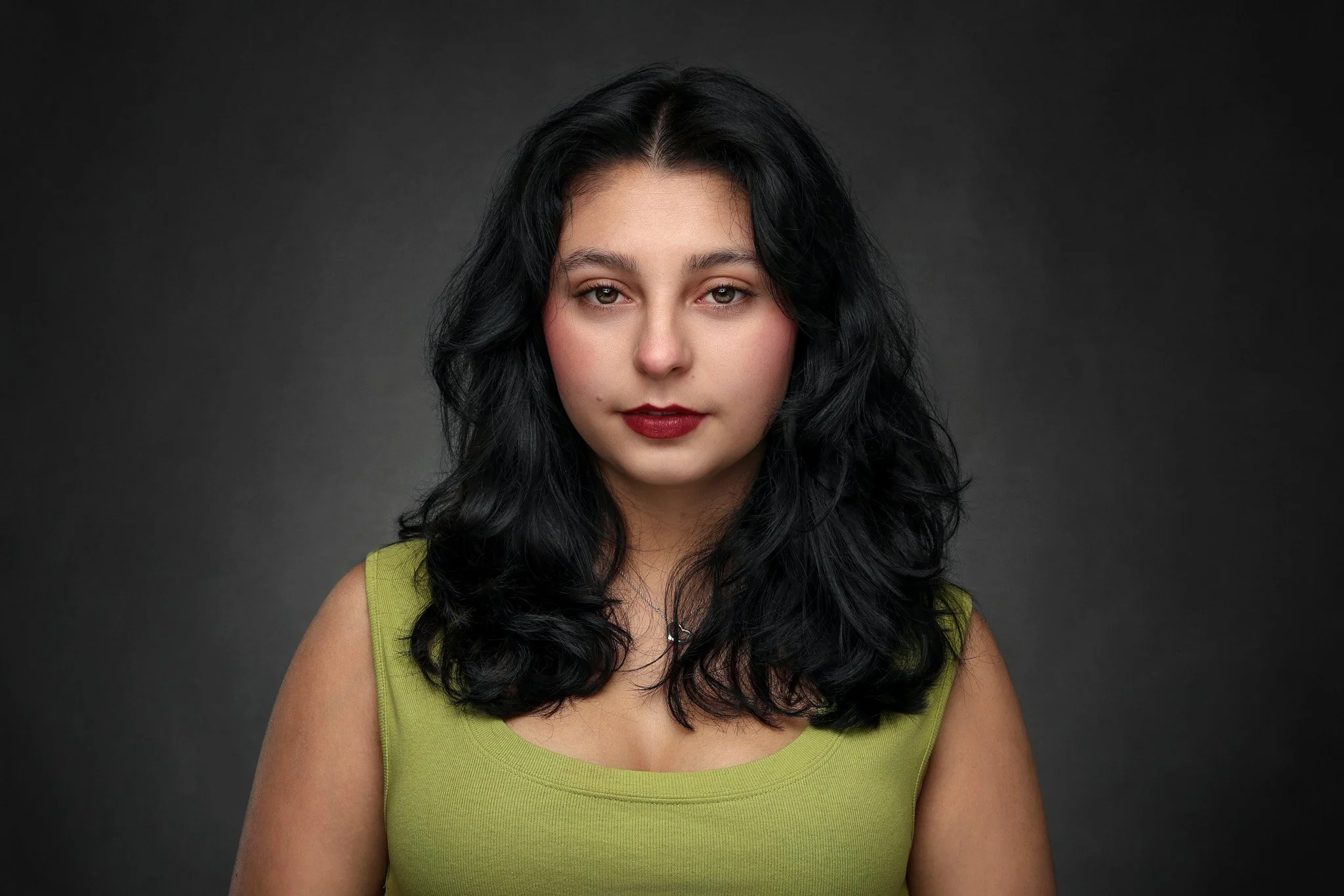 A young woman with long black hair, fair skin, and red lipstick gazing directly at the camera, wearing a sleeveless green top against a dark grey background.