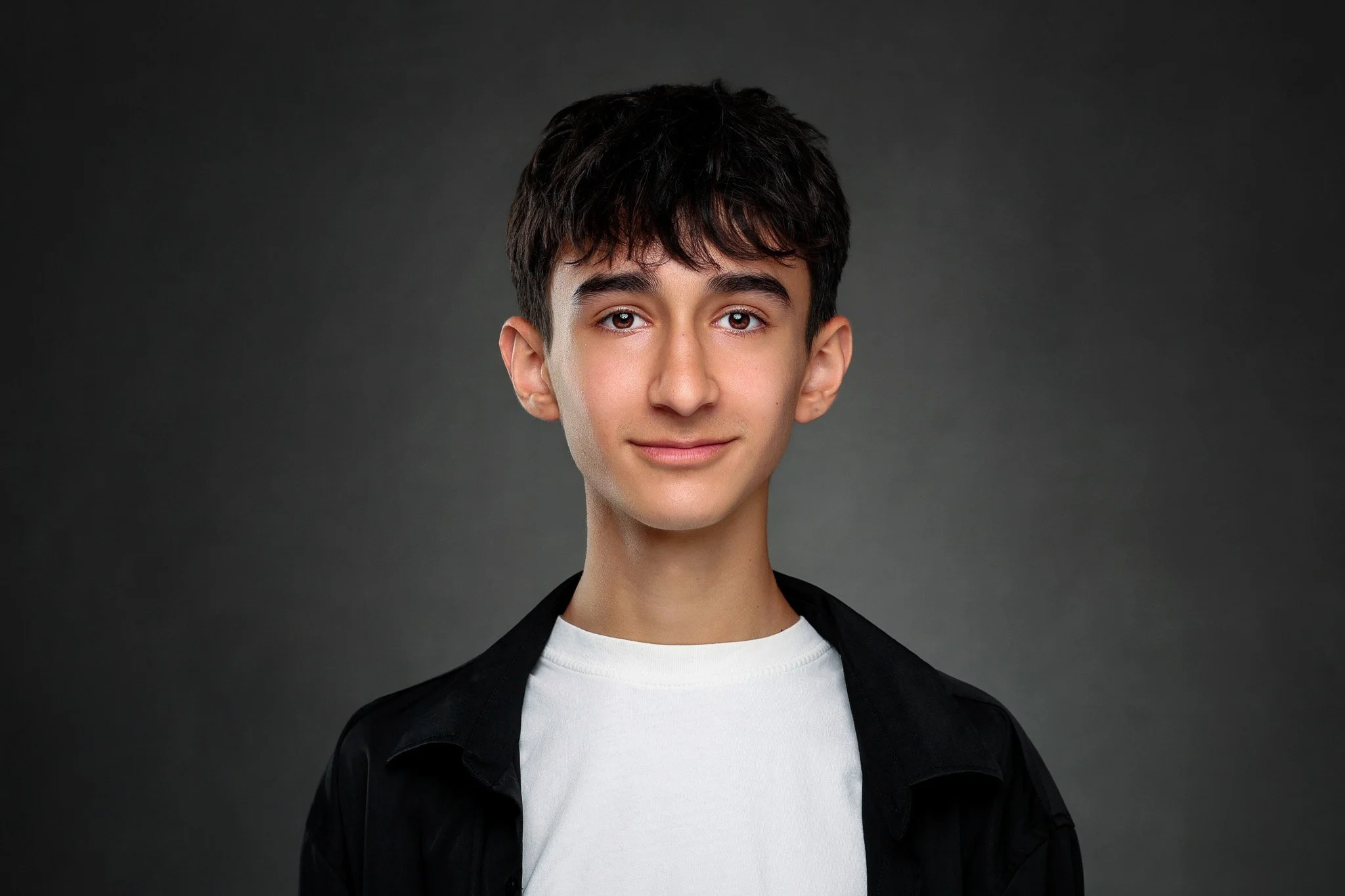 A young man with dark hair and brown eyes, wearing a white t-shirt and black jacket, against a dark grey background.
