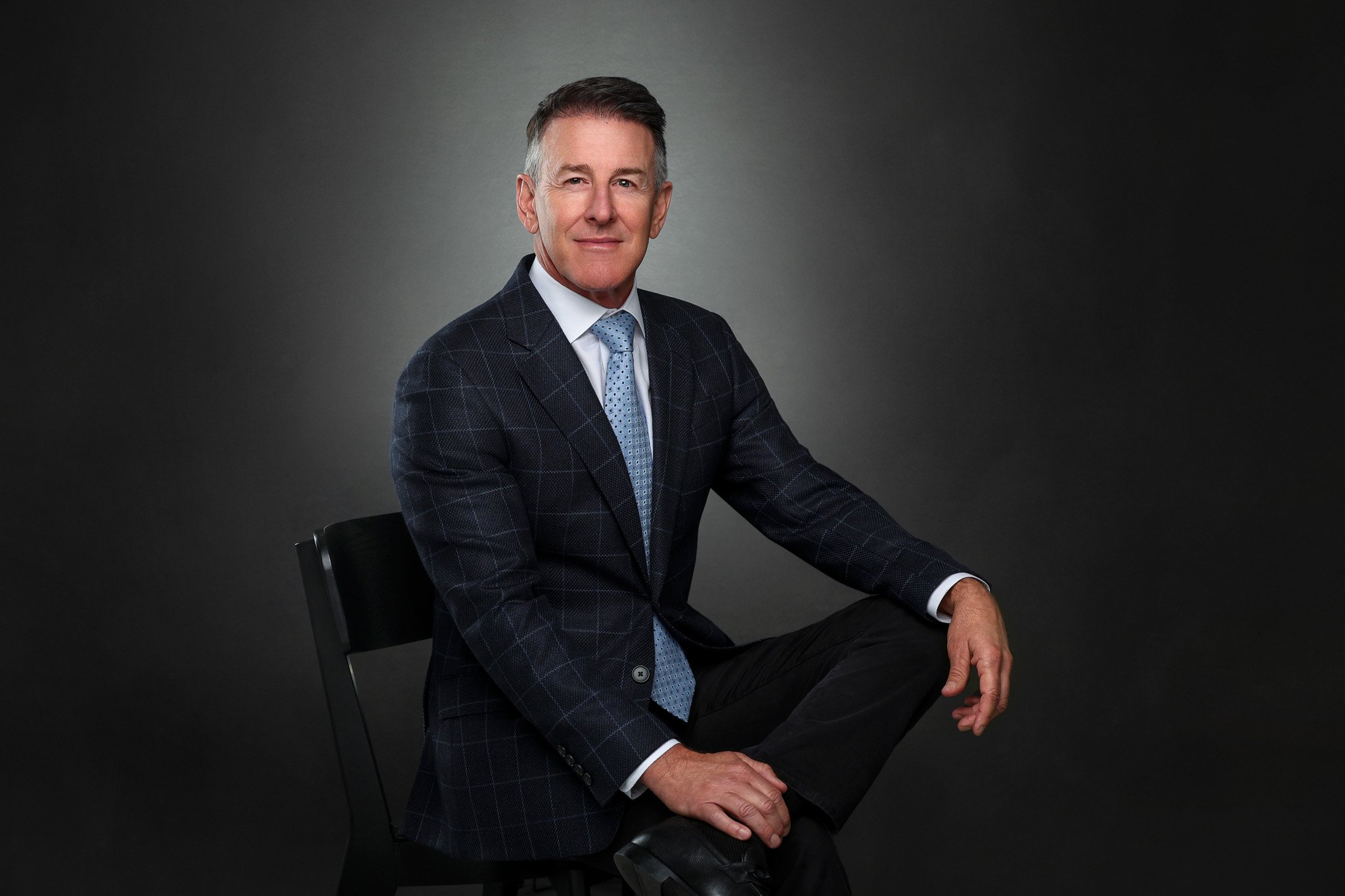 A middle-aged man in a dark checked suit, white shirt, and light blue tie, sitting on a black chair against a dark studio background.