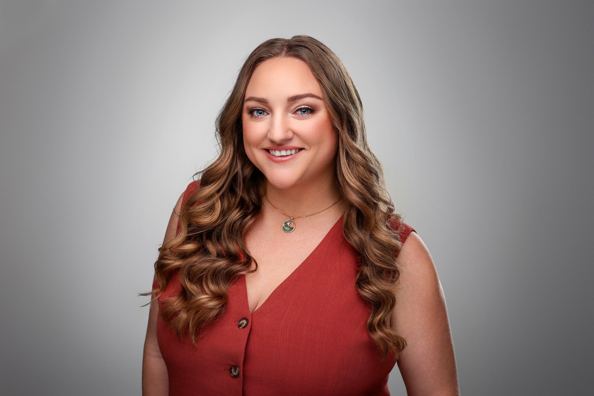 Portrait of a woman with long wavy brown hair, blue eyes, wearing a sleeveless rust-colored top with buttons, a gold necklace with a circular pendant, smiling against a plain gray background.