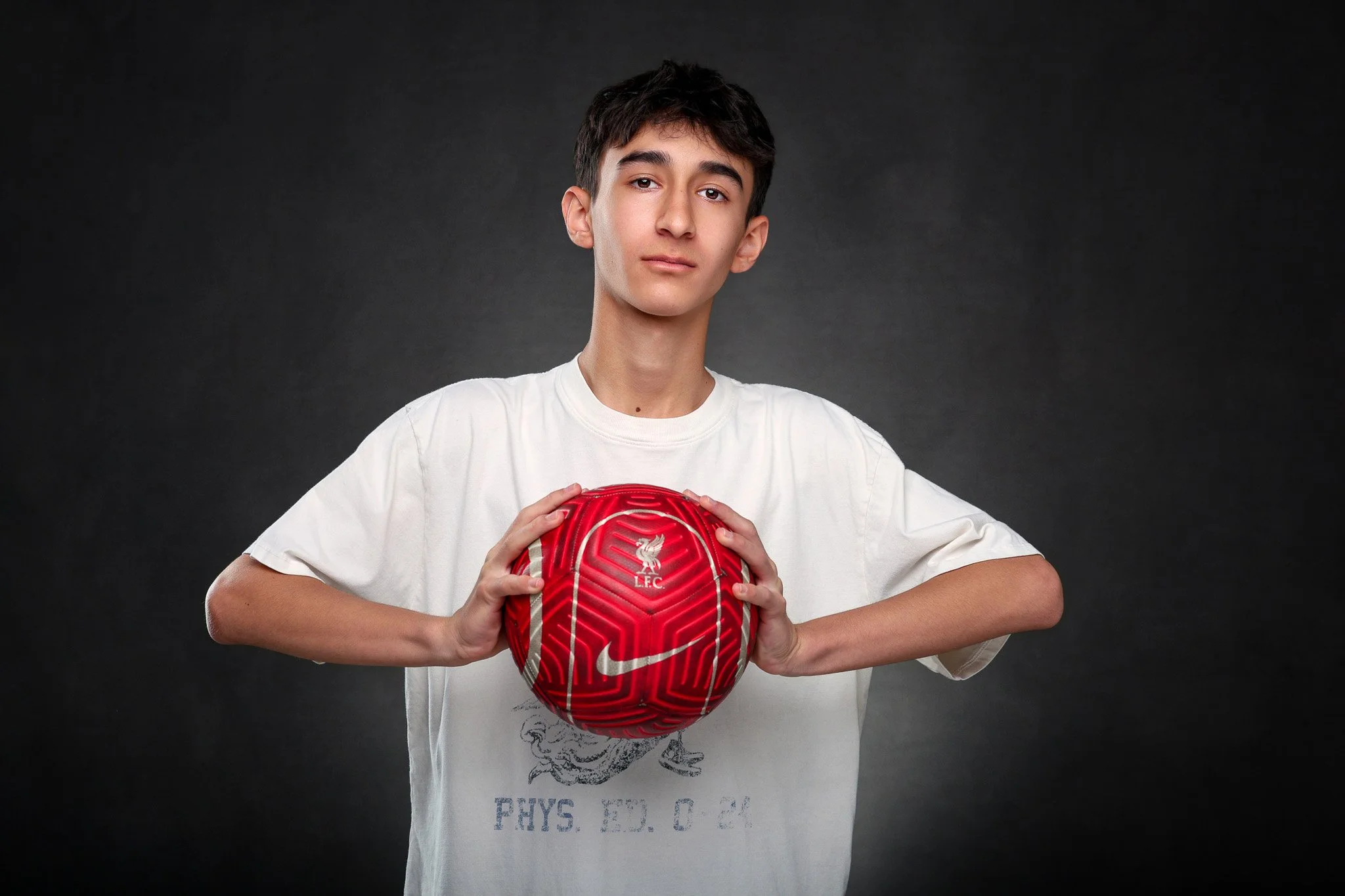 A young man holding a red Liverpool football club soccer ball against a dark gray background.