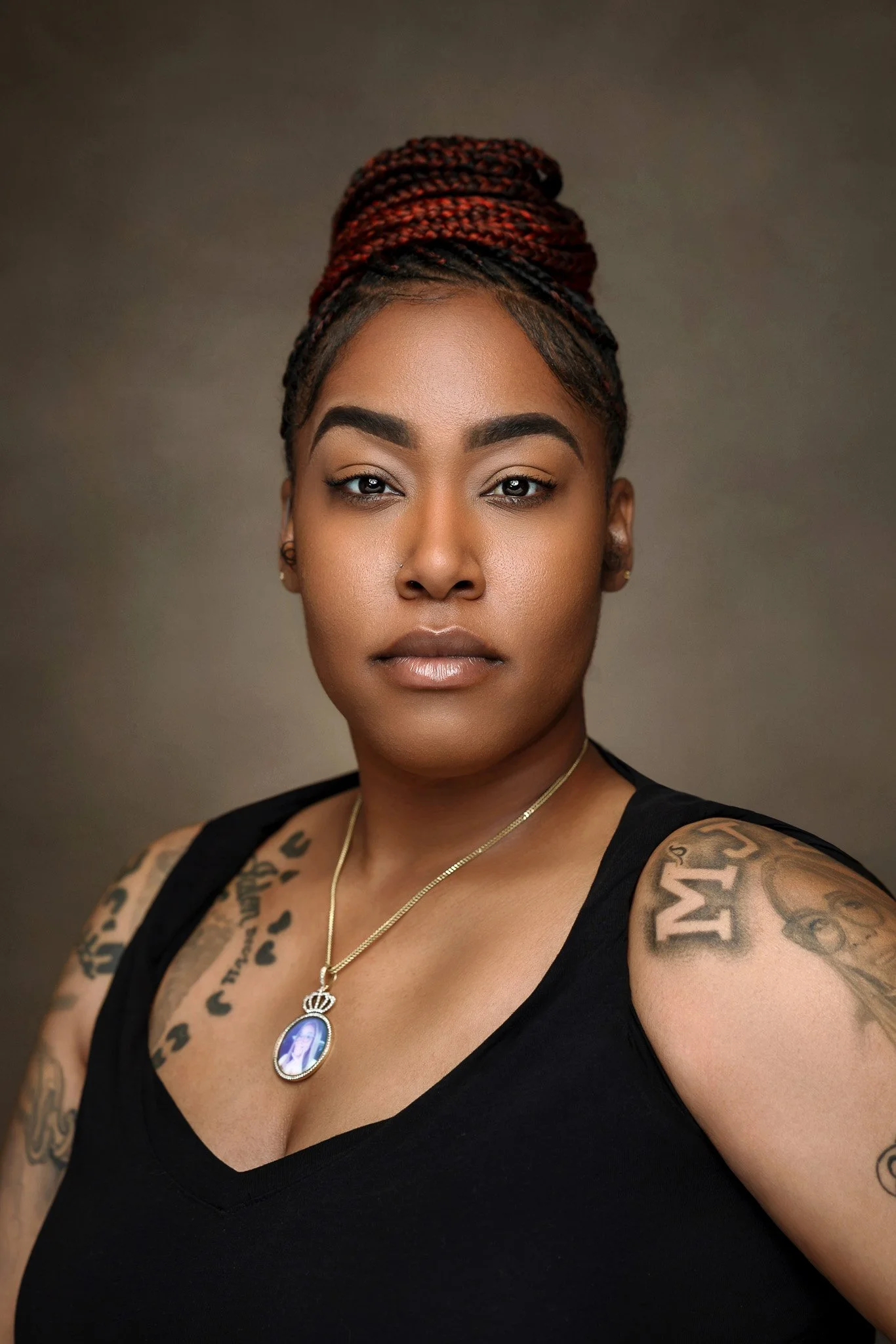A woman with braided hair updo, tattoos on shoulders and arms, wearing a black top, gold necklace, and a pendant with an image, posing against a neutral background.
