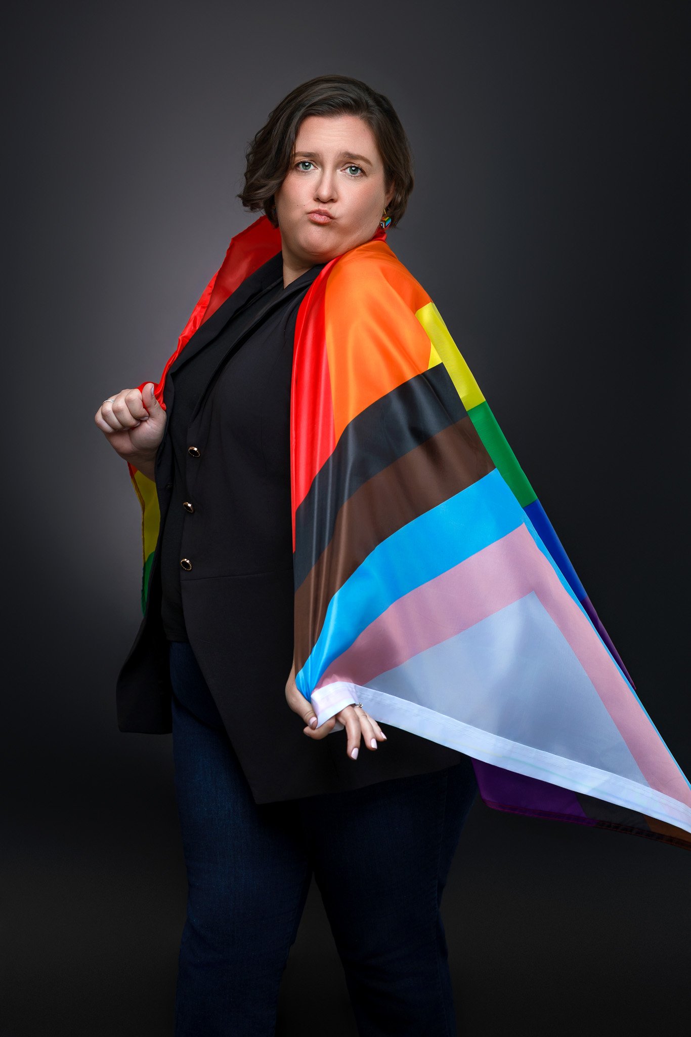 A woman with short brown hair is wearing a black blazer and jeans, holding a rainbow pride flag draped over her shoulders, standing against a dark background.