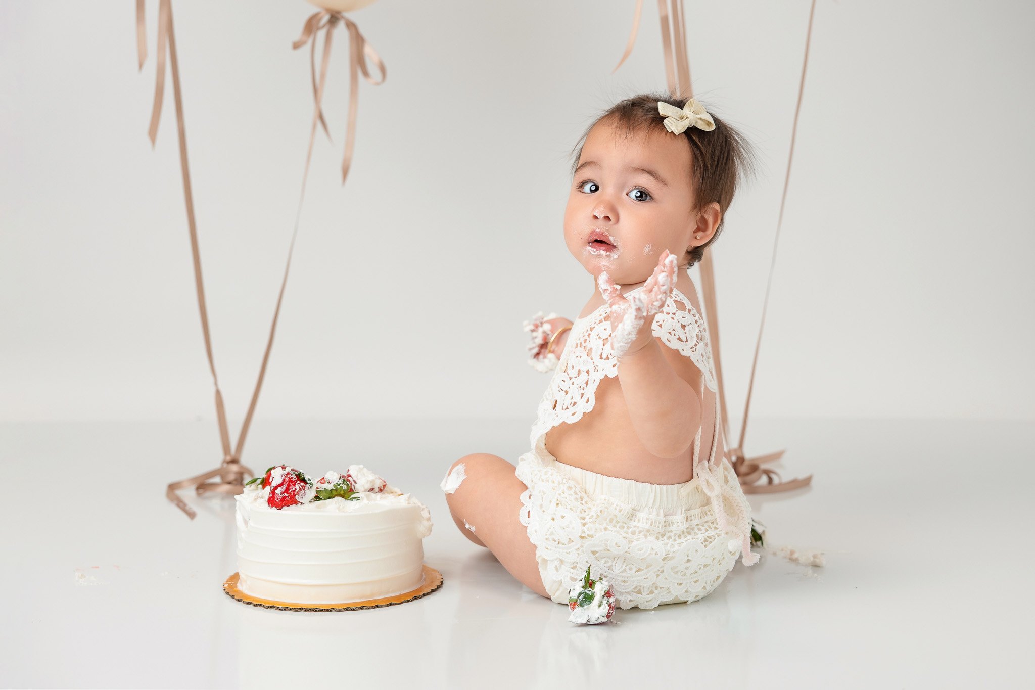 Baby sitting on the floor in front of a cake, wearing a lace outfit, with frosting on hands and face, surrounded by balloons.