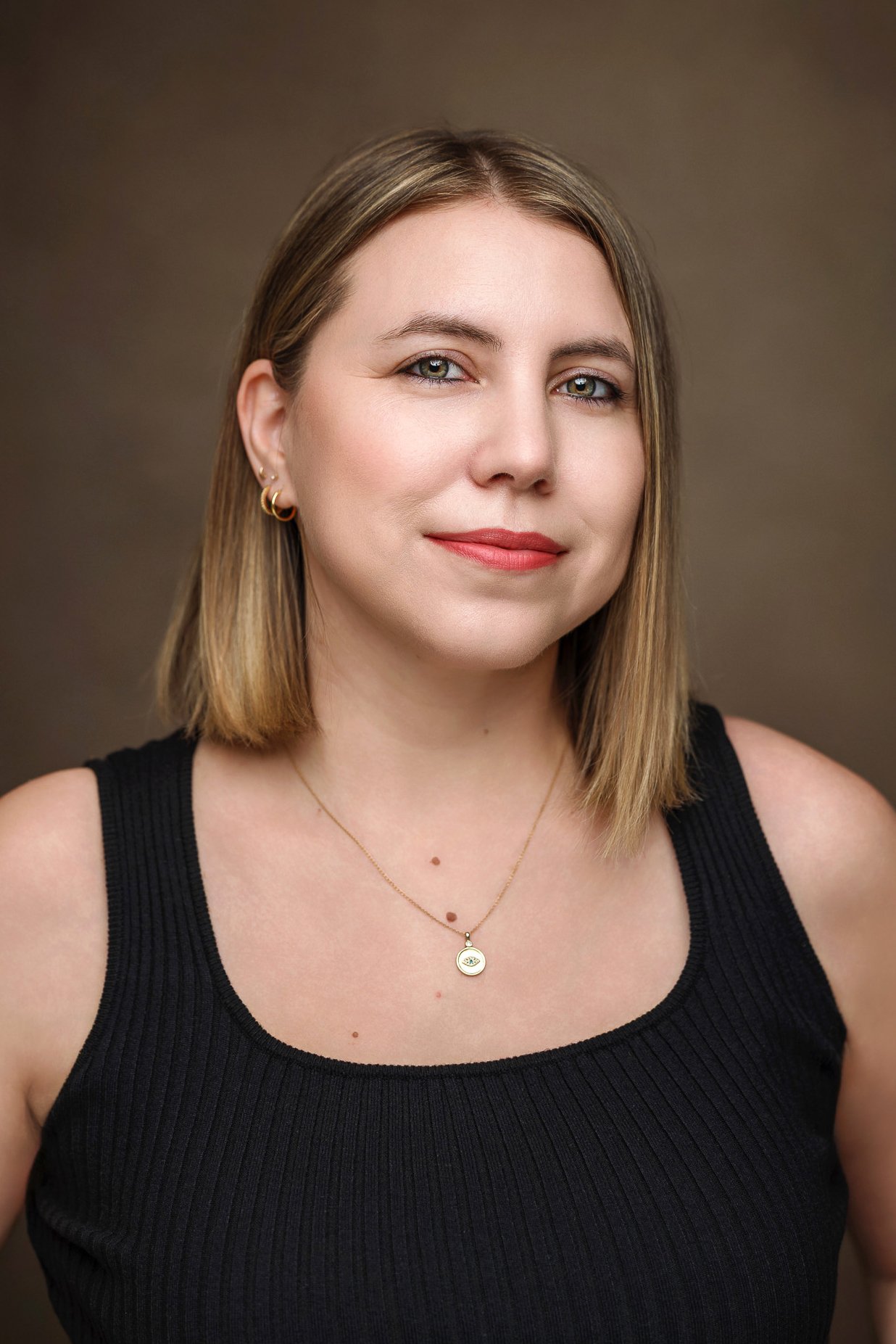 A woman with shoulder-length light brown hair, wearing gold hoop earrings, a gold necklace with a circular pendant, and a black sleeveless top, looking at the camera with a slight smile against a plain brown background.
