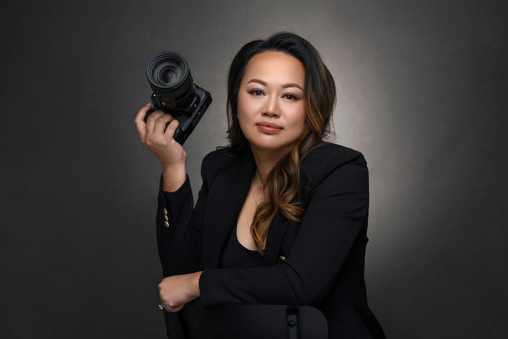 A woman with long wavy hair, wearing a black blazer, holding a professional camera, posing for a portrait against a neutral background.