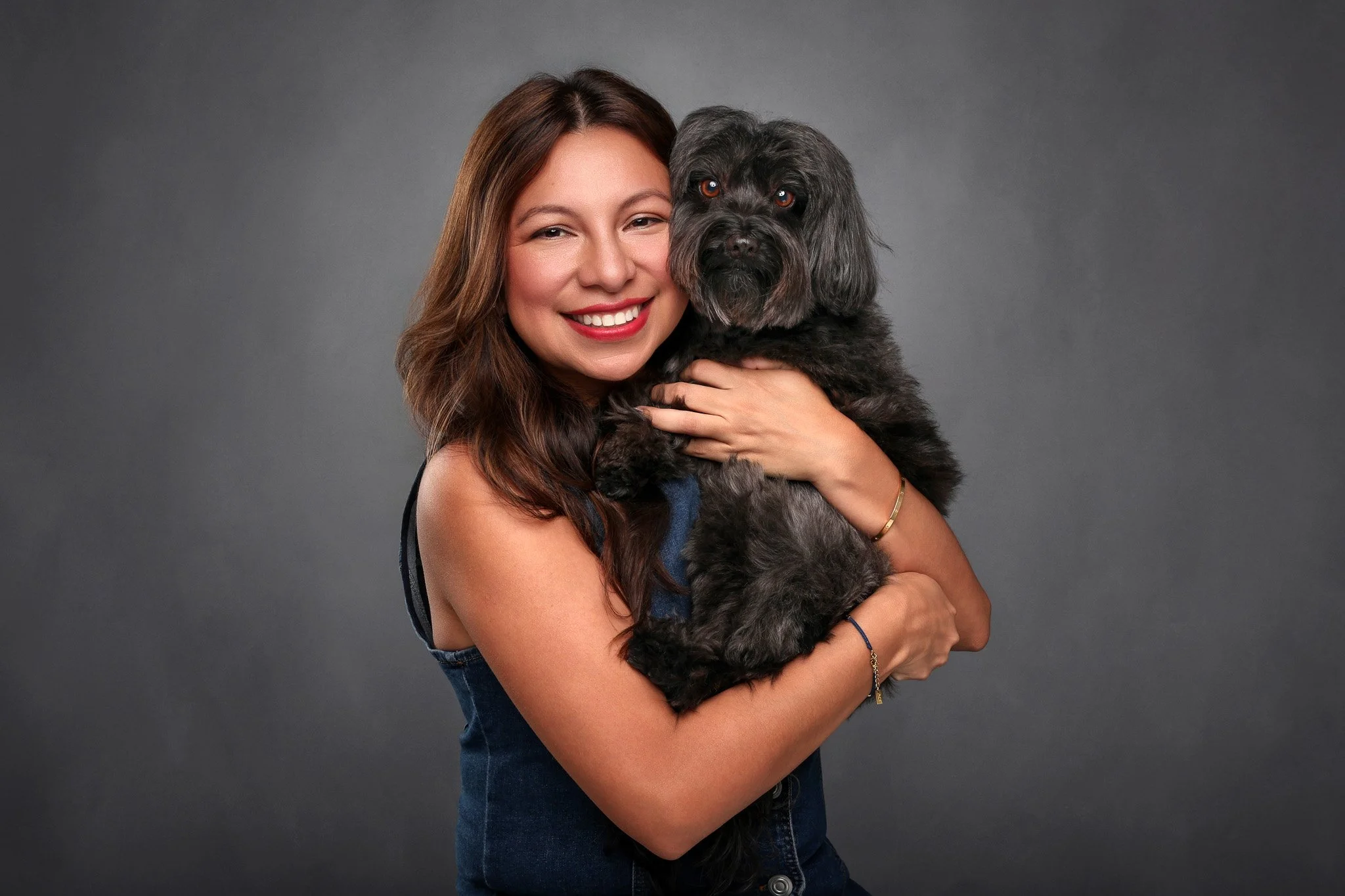 A woman with long brown hair smiling while holding a black and gray dog, both looking at the camera, against a gray background.