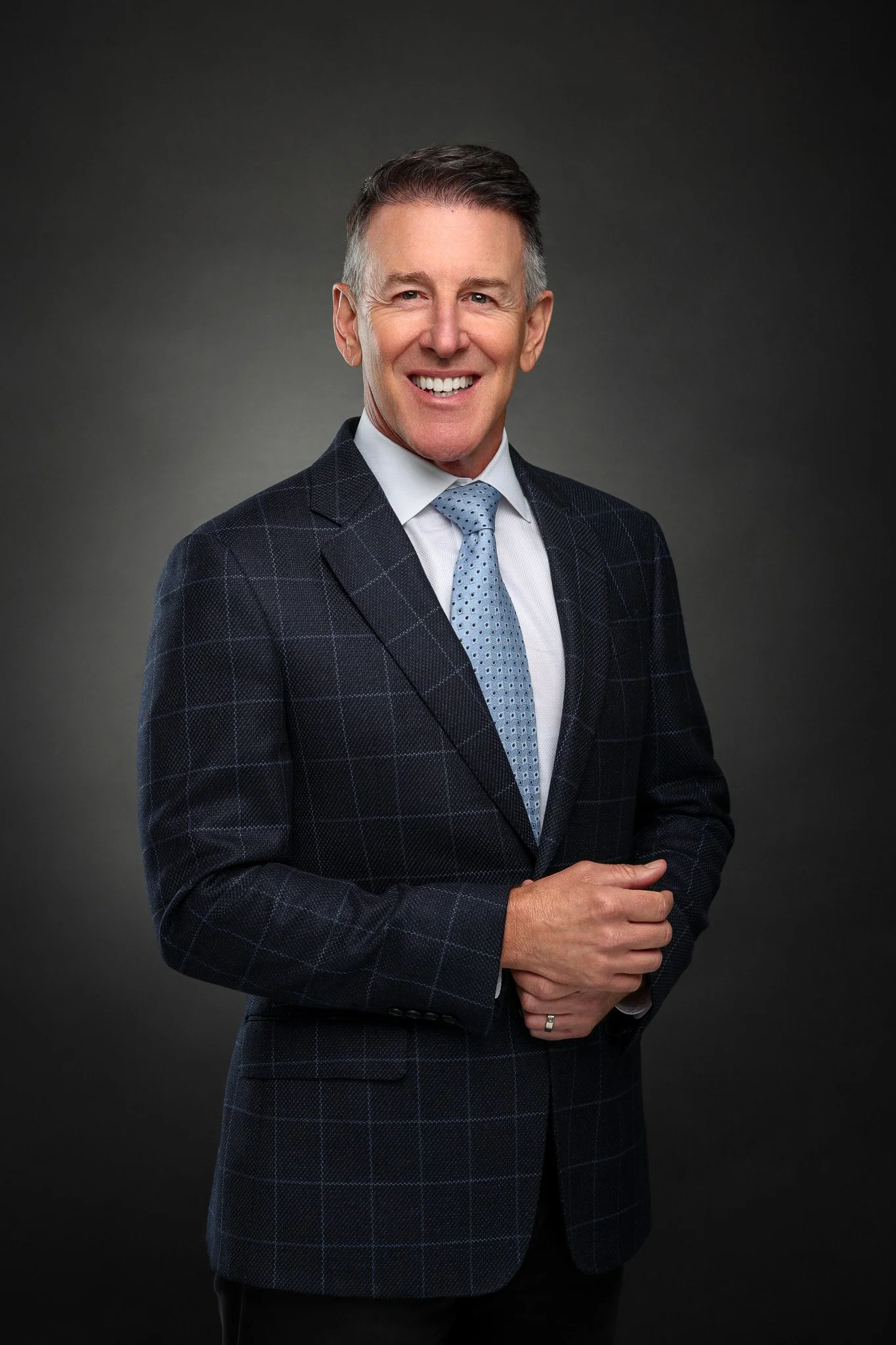 A man in a dark checkered suit with a light blue tie, smiling against a dark gray background.