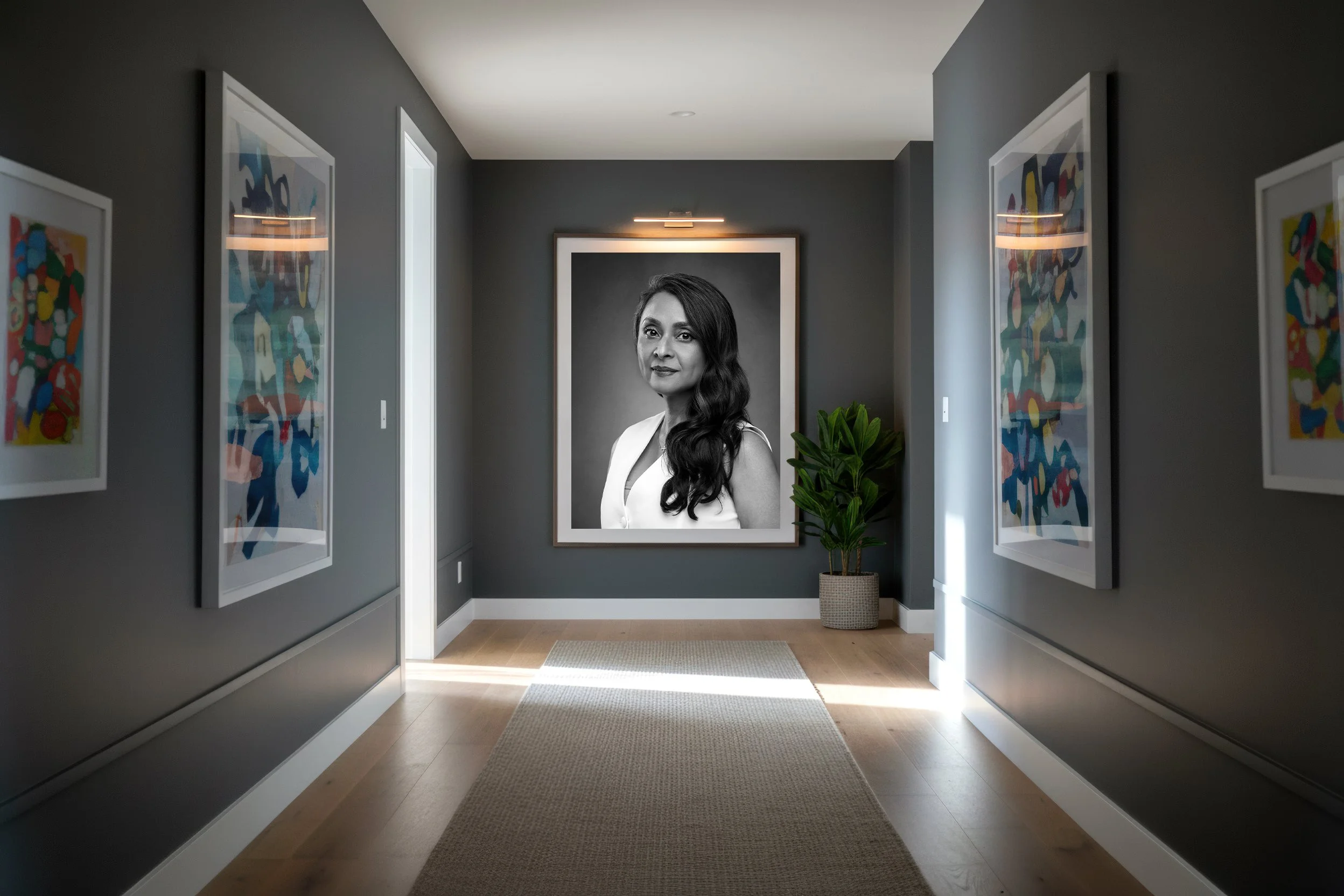 Gallery hallway with a large portrait of a woman with long dark hair, black and white, on a dark gray wall. Colorful abstract artwork framed on either side. Small potted plant to the right of the portrait, on a light wooden floor with a rug in front.