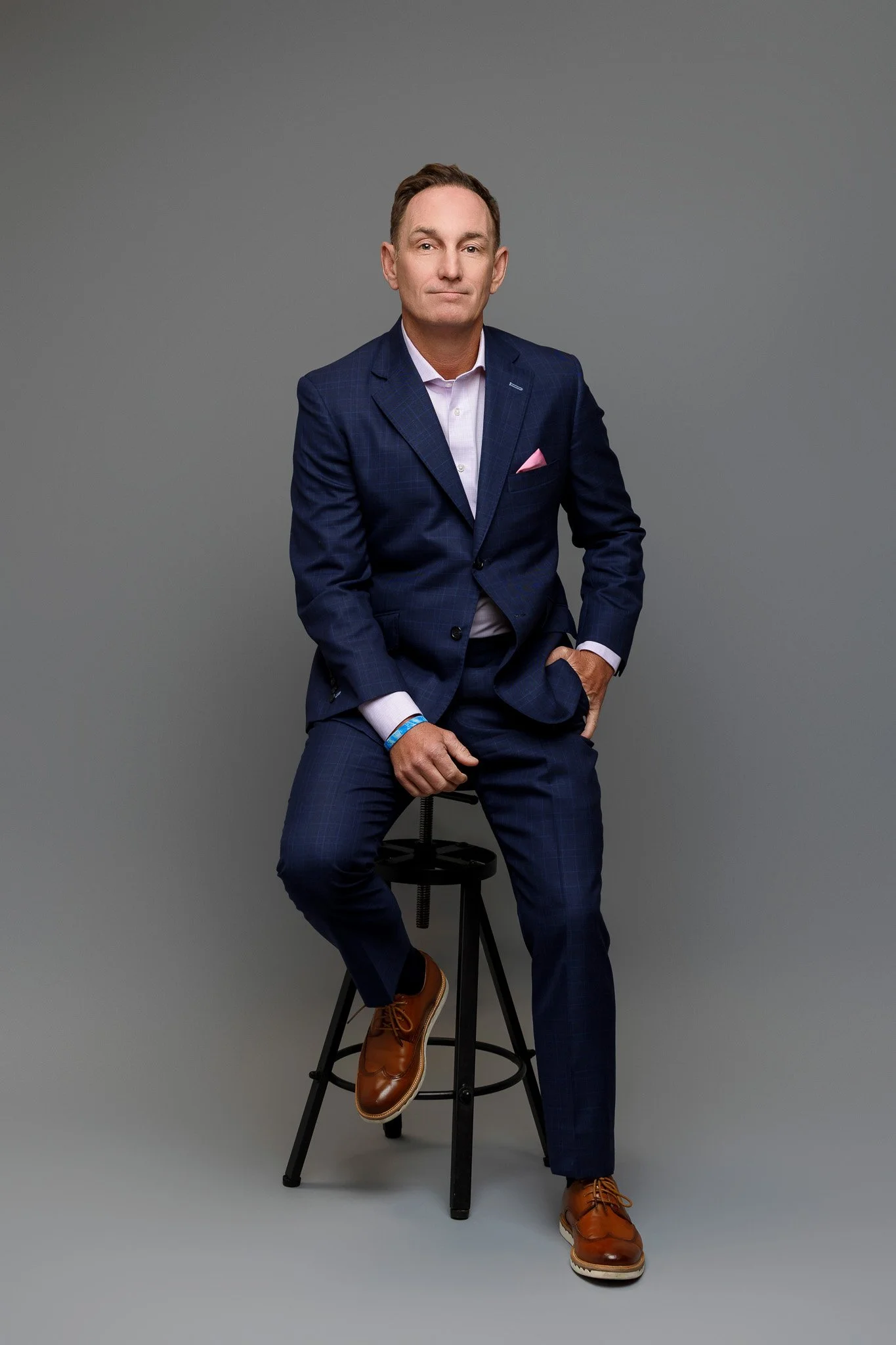 A man in a navy blue suit sitting on a black stool against a gray background.