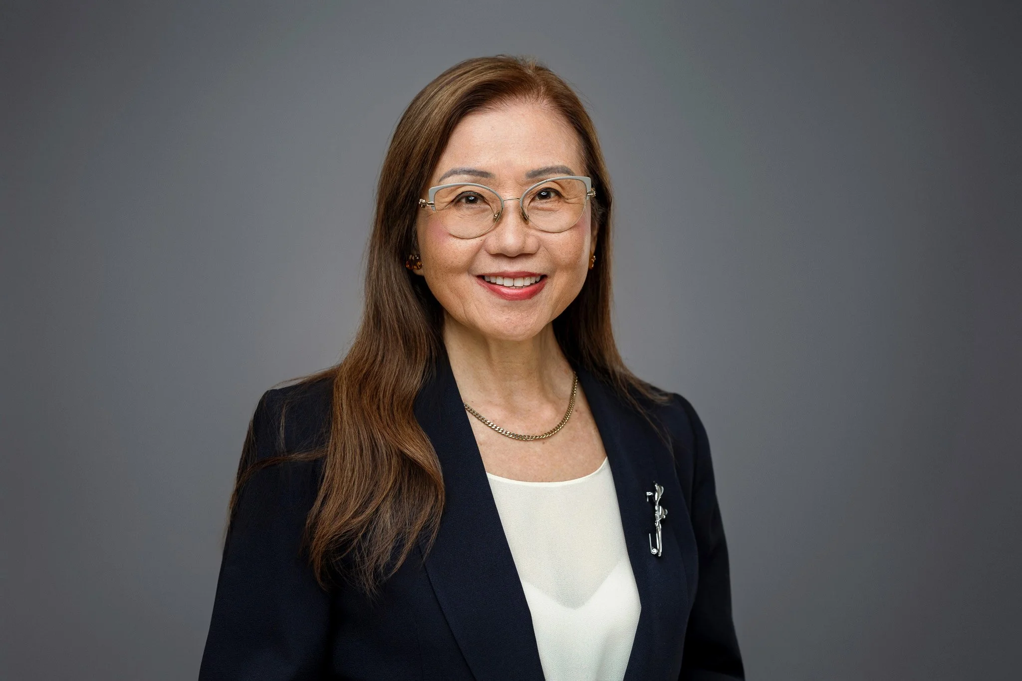 A professional woman with long brown hair, wearing glasses, a navy blazer with a brooch, and a white top, smiling at the camera against a gray background.