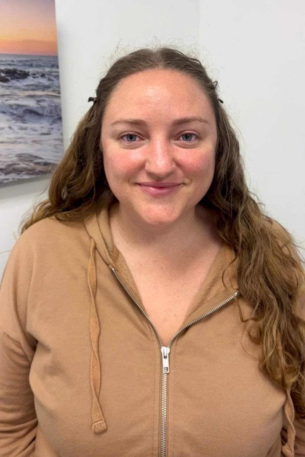 A woman with curly brown hair, blue eyes, and fair skin smiling at the camera, wearing a tan hoodie with a zipper, with a beach sunset photograph on the wall behind her.