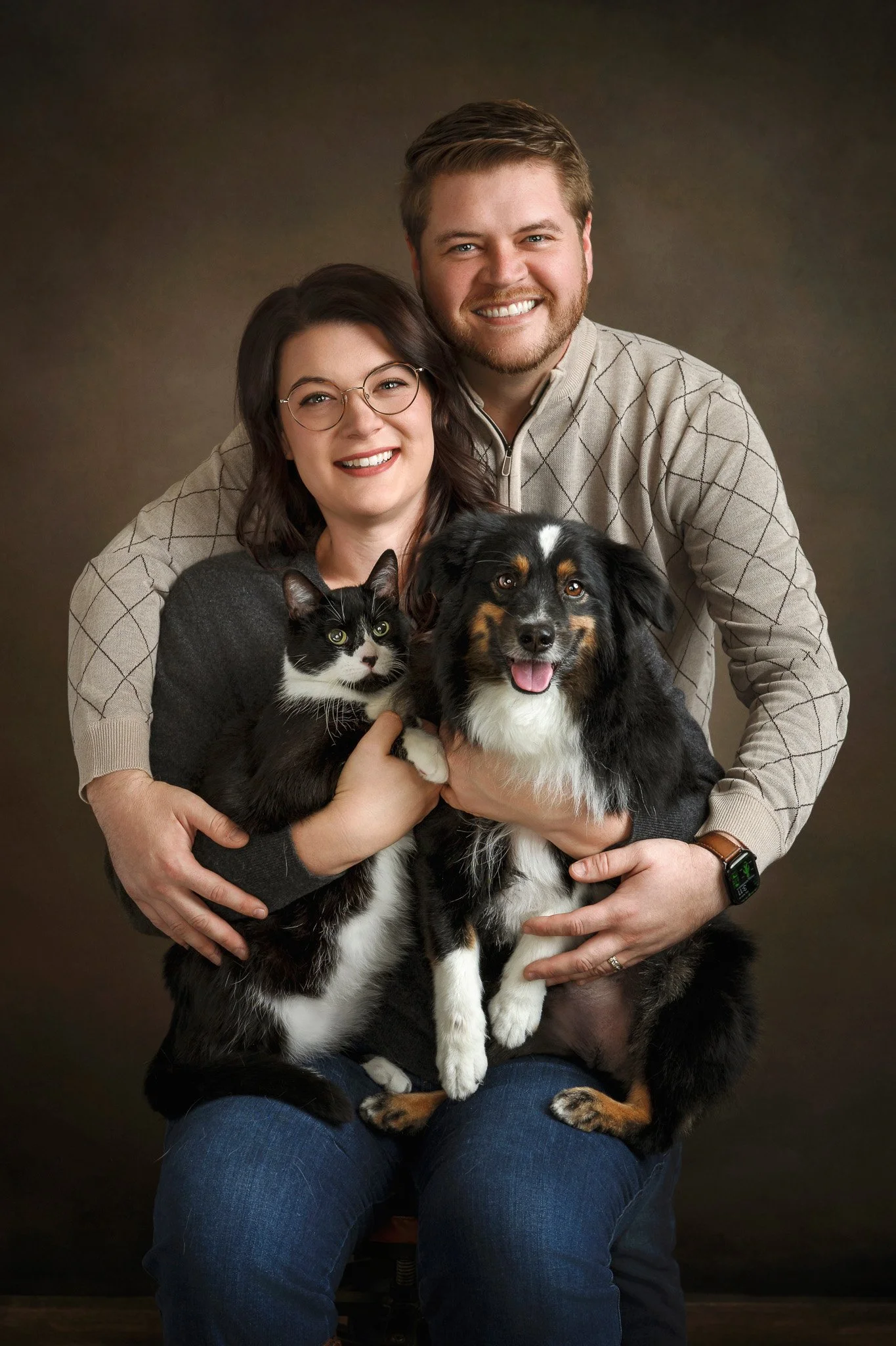 A smiling couple poses with their black and white cat and Australia shepherd dog in front of a dark background. The woman has shoulder-length dark hair, glasses, and wears a gray sweater. The man has short light brown hair, a beard, and wears a beige
