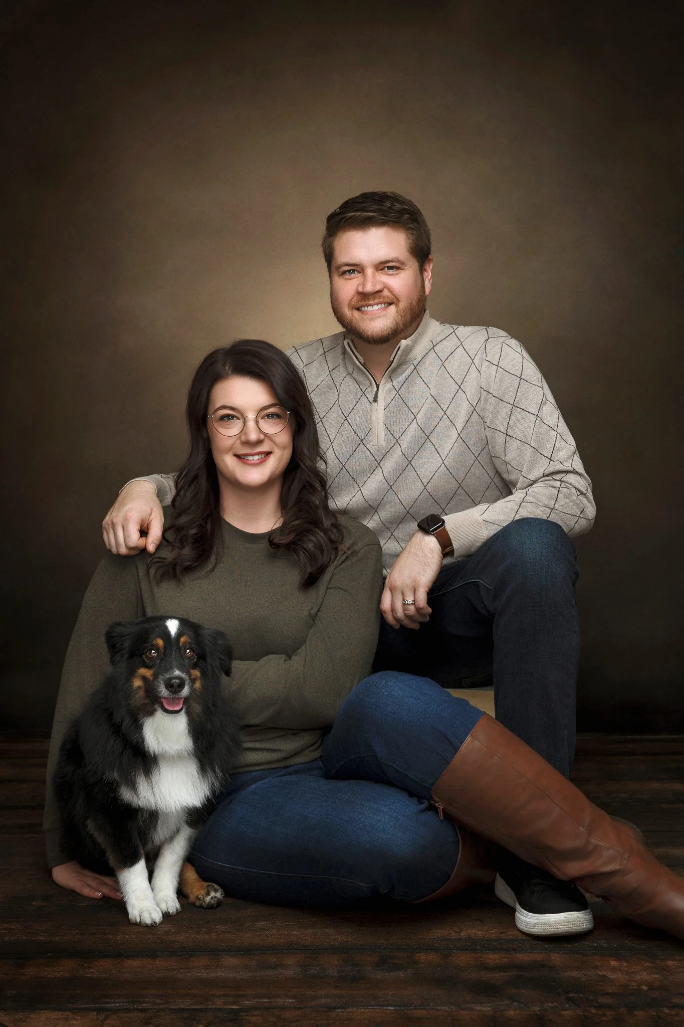 A smiling couple with their Australian Shepherd dog, sitting on a wooden floor against a dark brown backdrop.