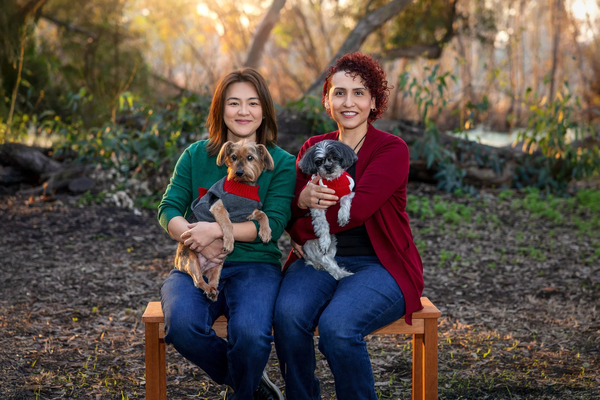 Two women sitting outdoors on a wooden bench, each holding a small dog dressed in sweaters, with trees and a river in the background during autumn.