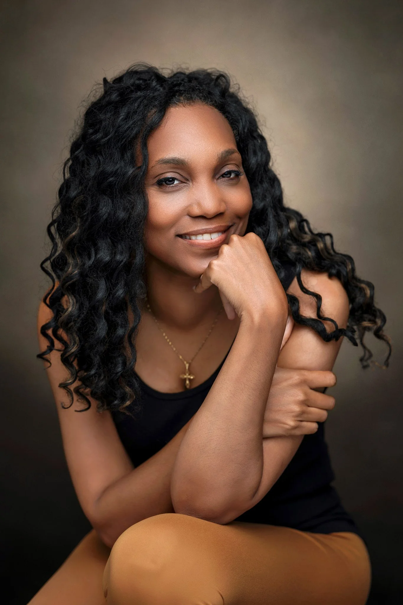 A woman with long, curly black hair, wearing a black top and a gold cross necklace, faces forward with a gentle smile, her chin resting on her hand, against a neutral background.