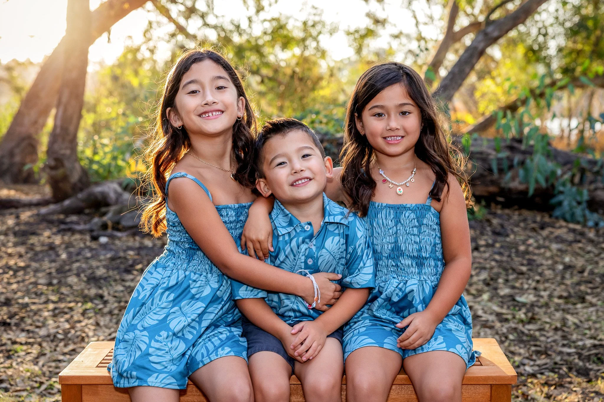 Three children, two girls and one boy, sitting on a wooden bench outdoors with trees and sunlight in the background. The children are smiling and wearing matching blue patterned outfits.