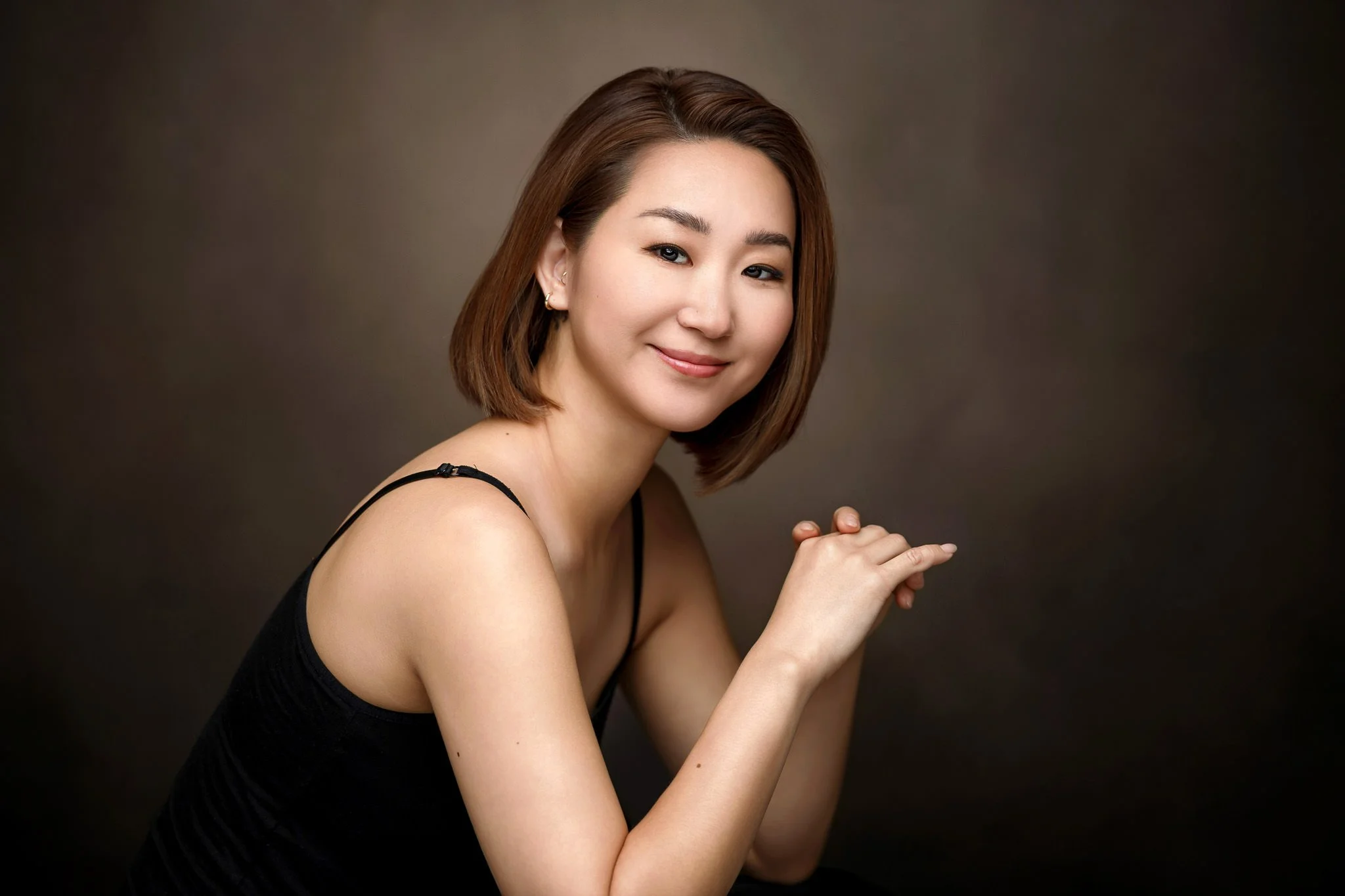 A woman with short brown hair, wearing a black sleeveless top, is smiling gently with her hands clasped together against a brown gradient background.
