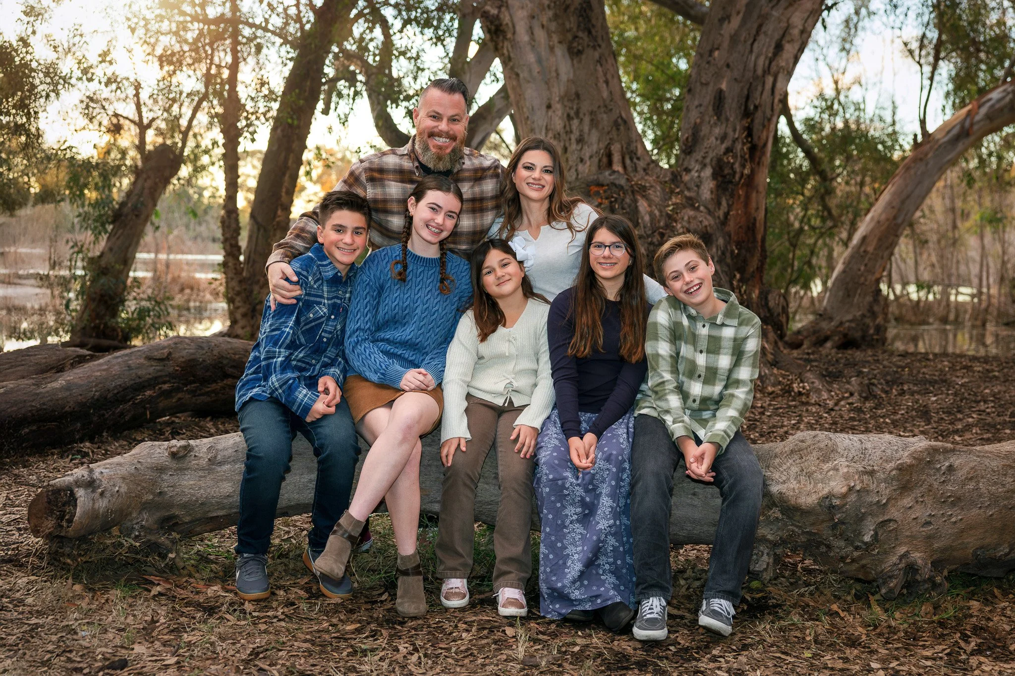 A family of eight, including children and adults, sitting on a log outdoors in a wooded area during sunset, smiling at the camera.