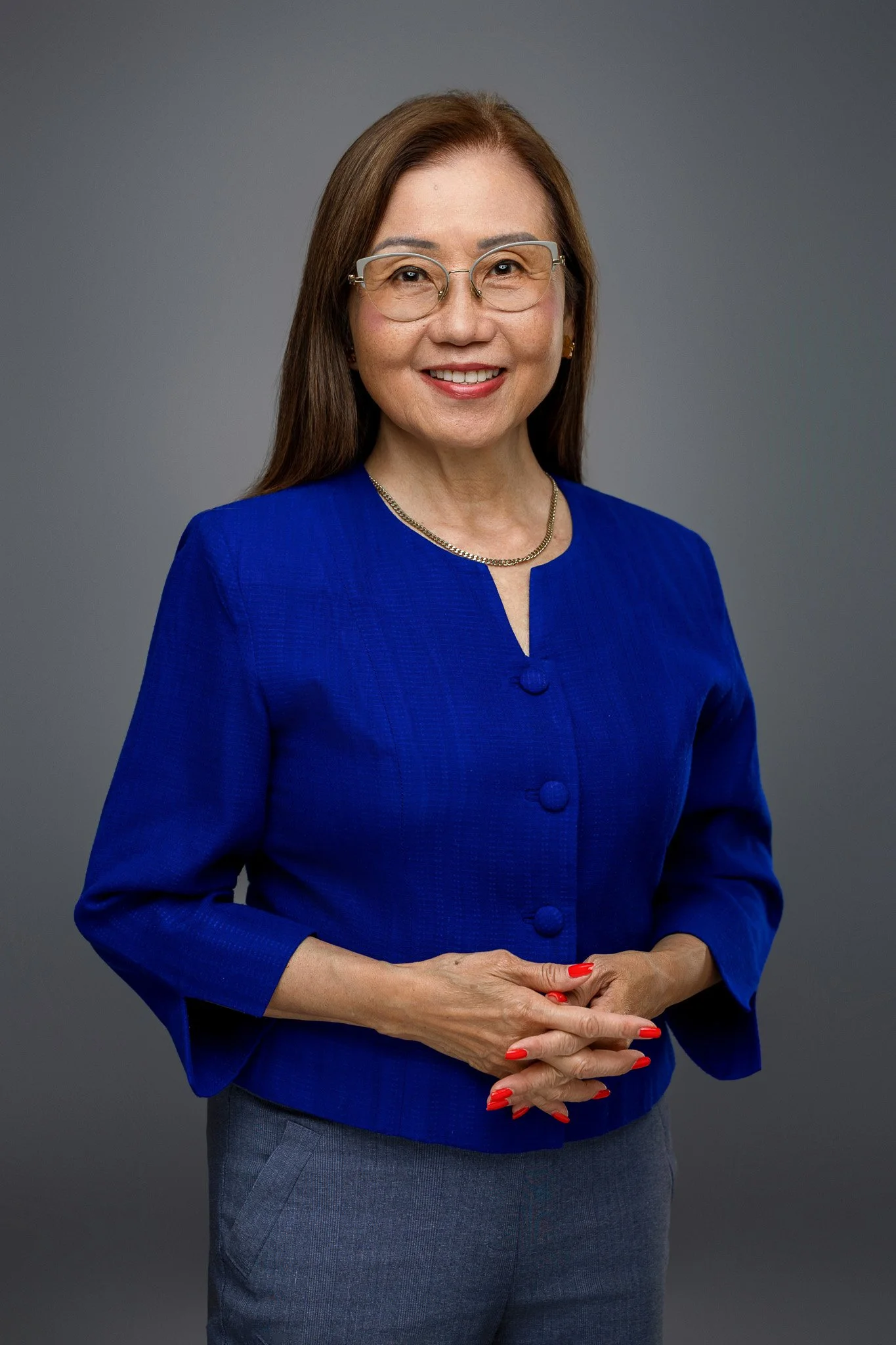A woman with shoulder-length brown hair, glasses, and red nail polish, smiling in a blue blazer and gray pants against a gray background.