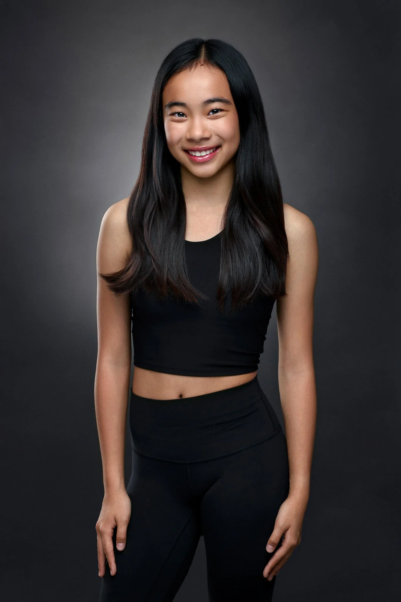 A young woman with long black hair smiling, wearing a black sleeveless top and black leggings, standing against a dark gray background.