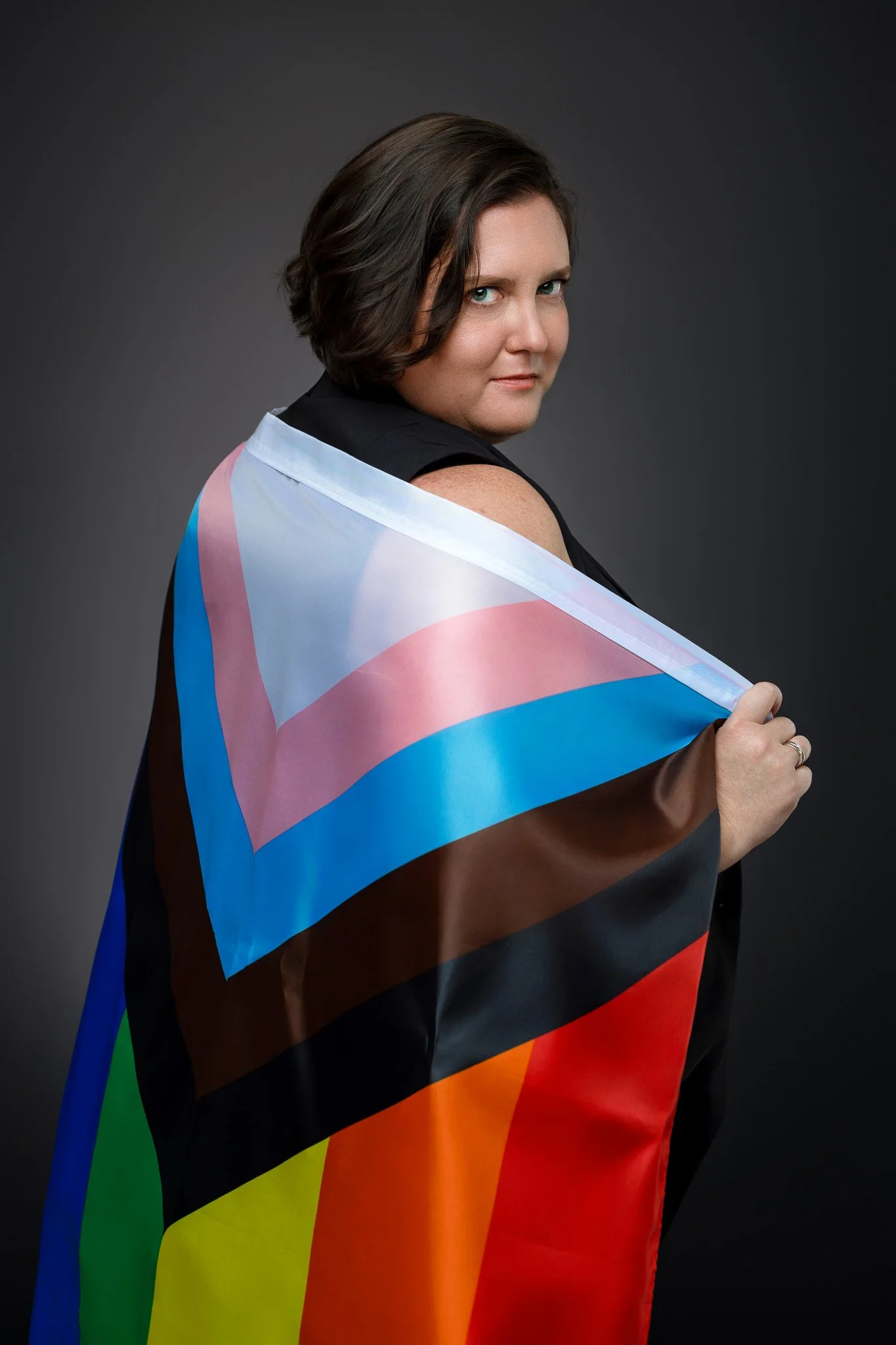 A woman with short dark hair looking at the camera, holding a transgender pride flag draped over her shoulder, against a dark background.
