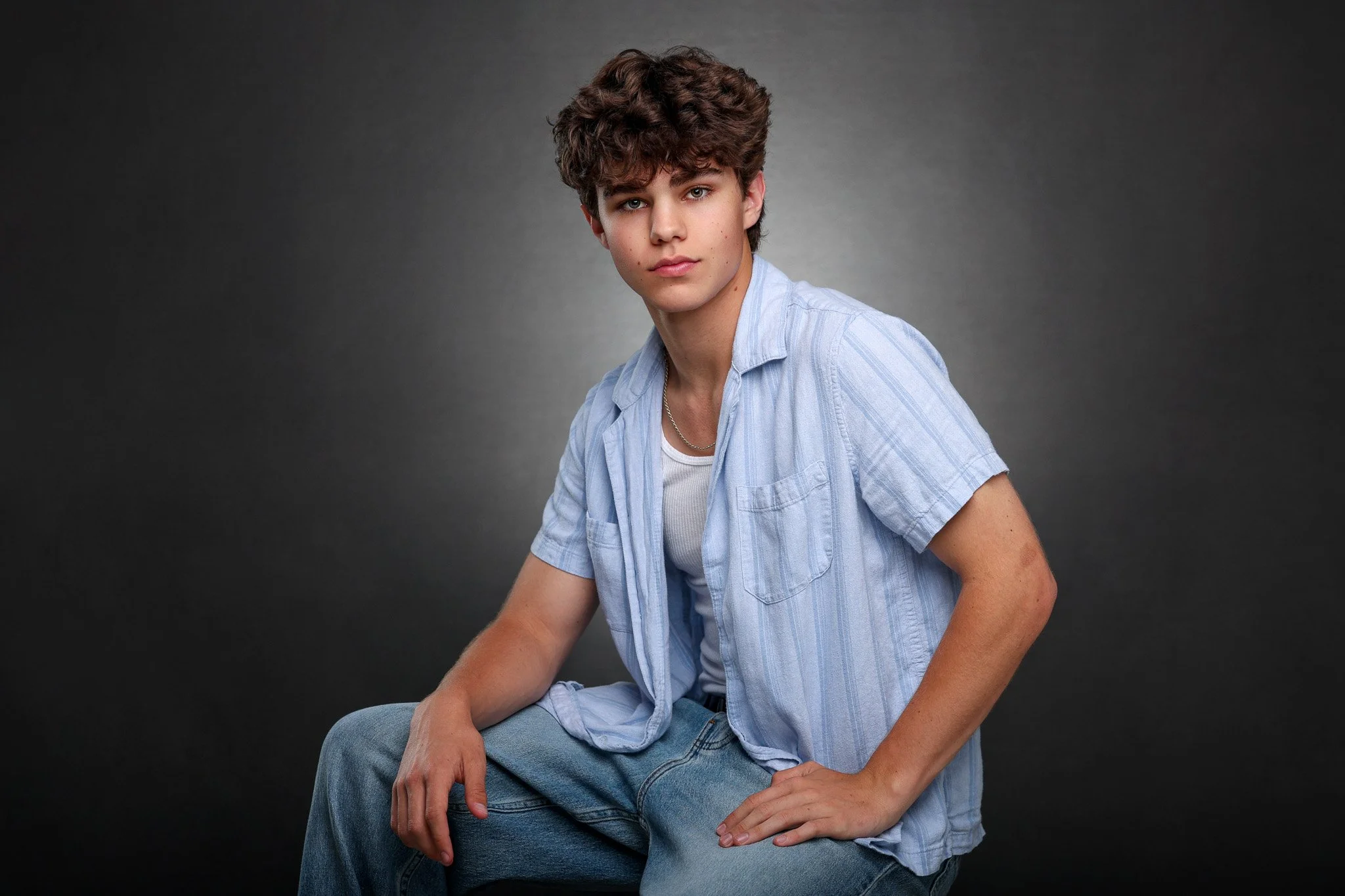 Young man with curly brown hair wearing a light blue shirt and jeans, sitting against a dark gray background.