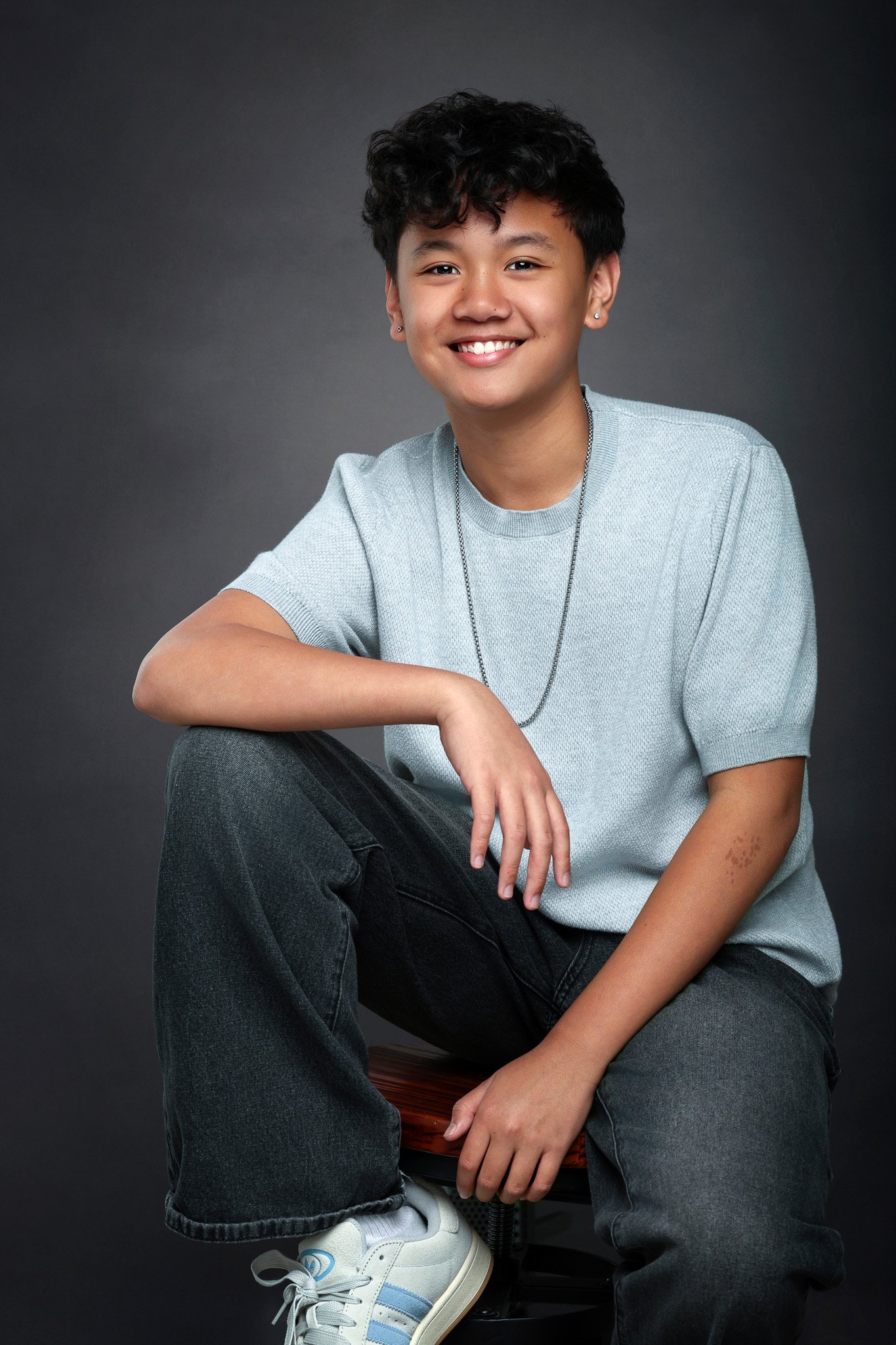 A young person with short, curly hair and earrings, sitting on a wooden stool against a dark background, smiling at the camera, wearing a light gray T-shirt, black jeans, and white sneakers.