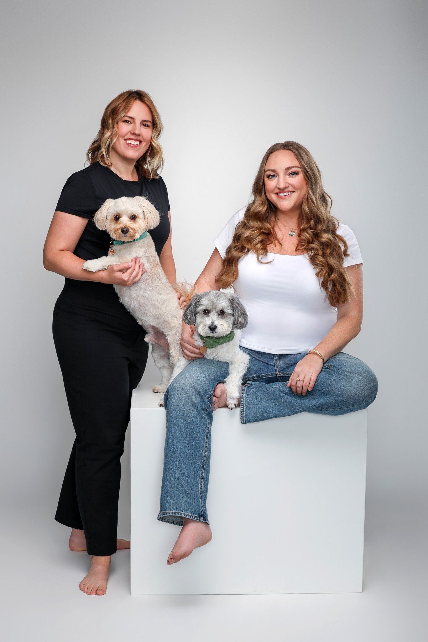 Two women sitting and standing, holding two small dogs, in a studio with a plain background.