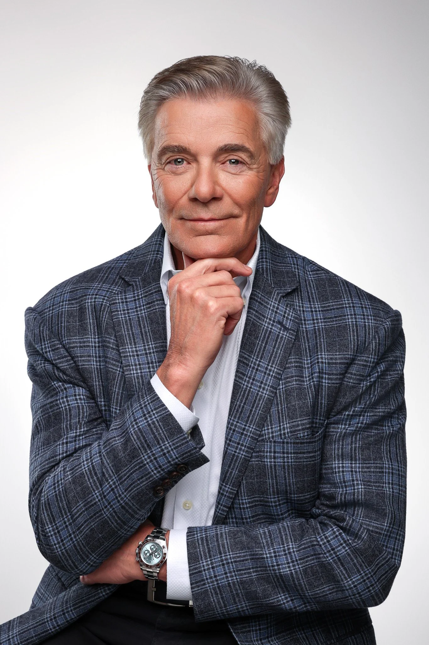 A confident older man with silver hair, dressed in a plaid blazer and white shirt, posing with one hand on his chin and the other resting on his arm, against a plain white background.