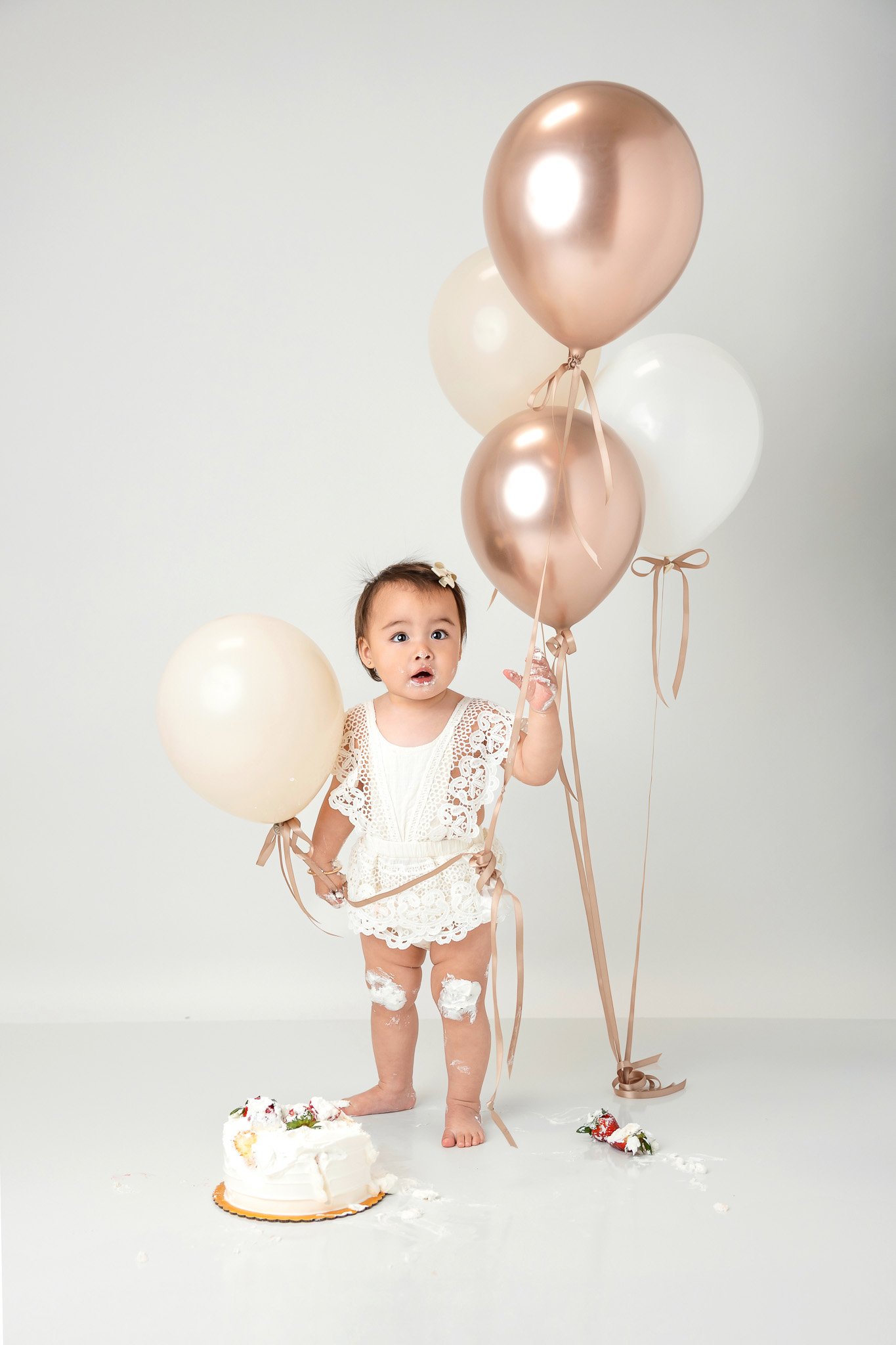 Baby in white lace outfit holding beige and white balloons, standing next to a partially eaten cake.