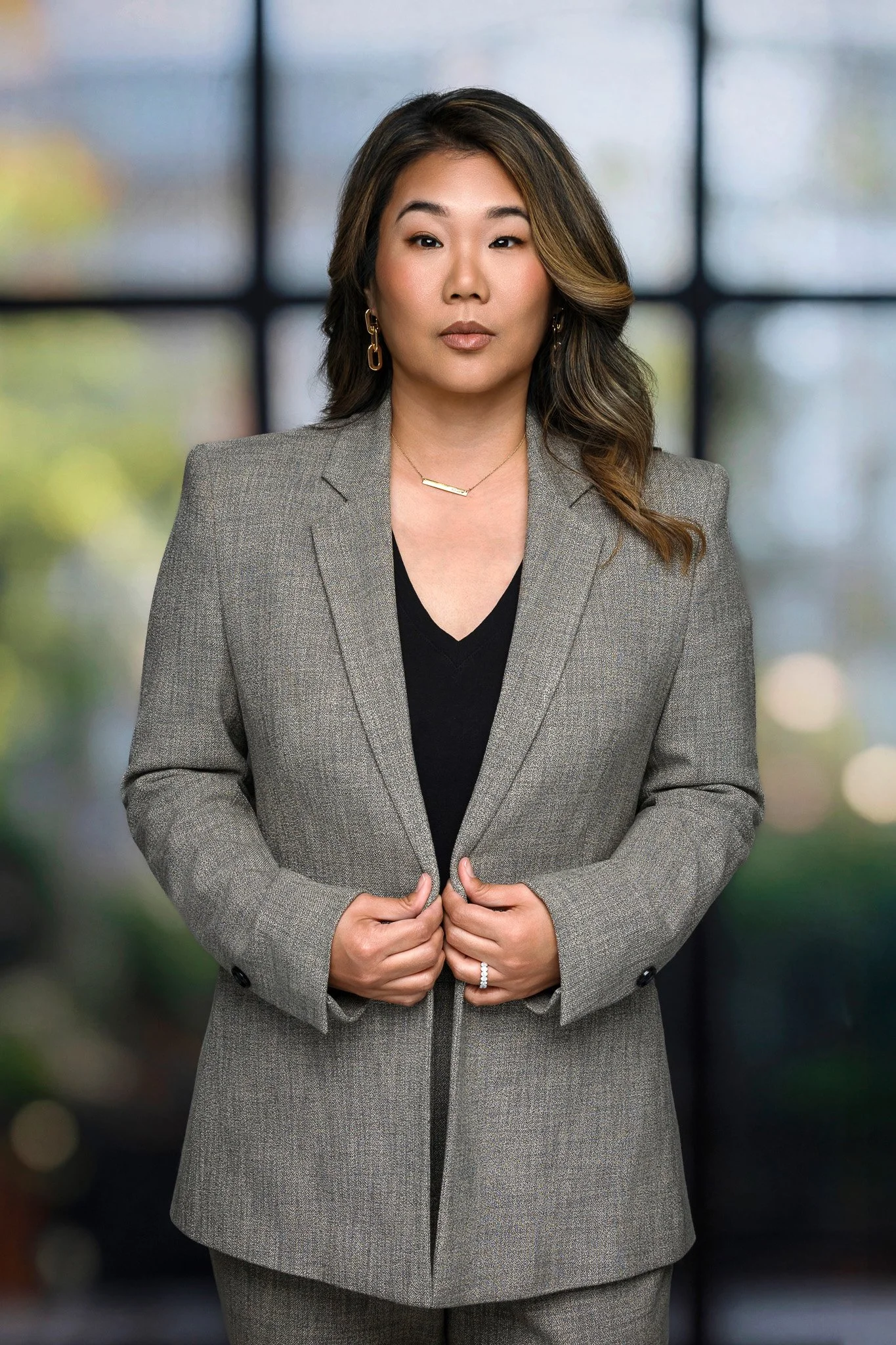 A professional woman with shoulder-length hair wearing a grey blazer over a black top, standing indoors with a blurred window background.