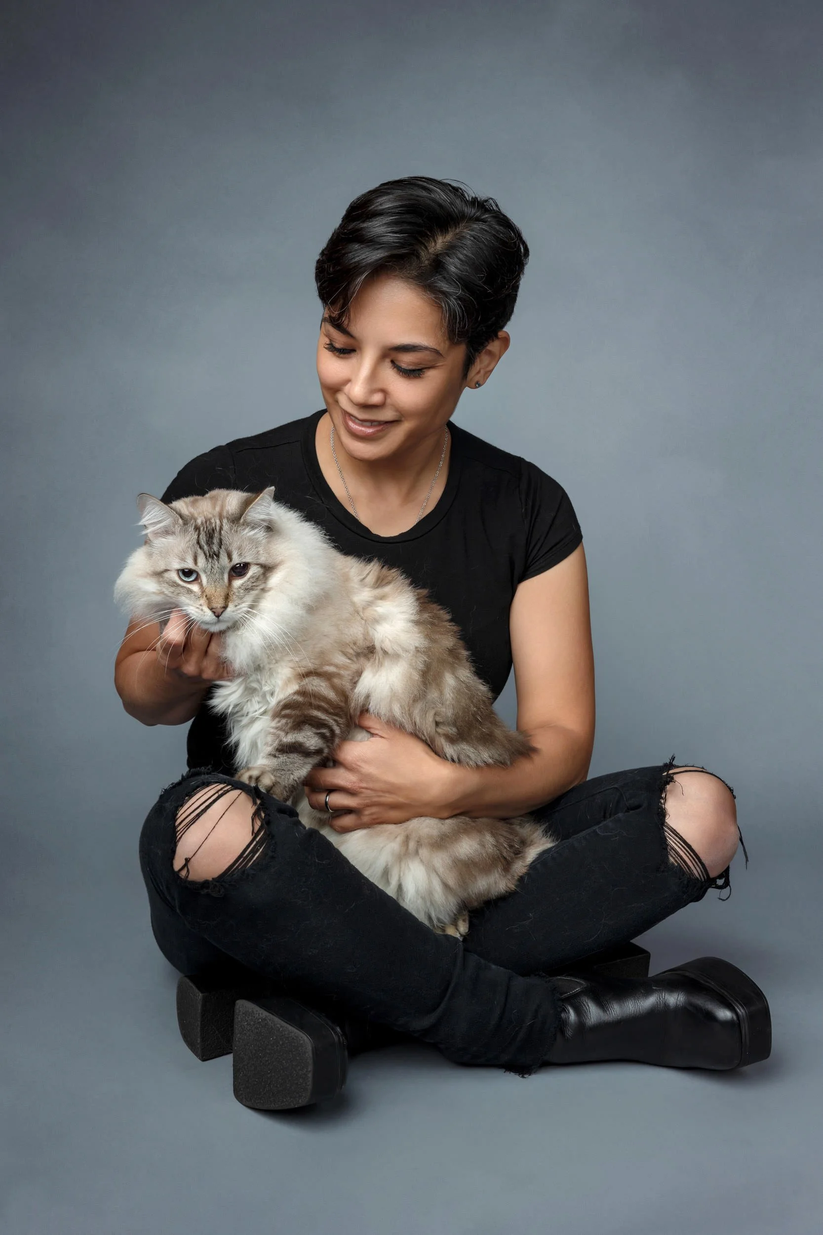 A woman with short black hair, wearing a black t-shirt and ripped black jeans, sitting cross-legged on a black block, holding a fluffy, light-colored cat with striking blue eyes against a plain gray background.